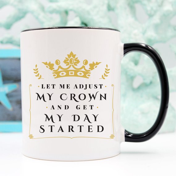 A humorous coffee mug with the phrase 'Let Me Adjust My Crown And Get My Day Started' printed on it, showcasing a fun and motivational design.