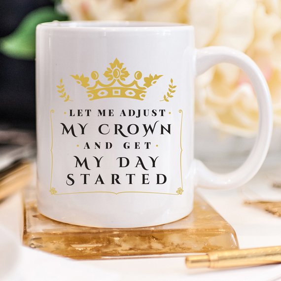 A humorous coffee mug with the phrase 'Let Me Adjust My Crown And Get My Day Started' printed on it, showcasing a fun and motivational design.