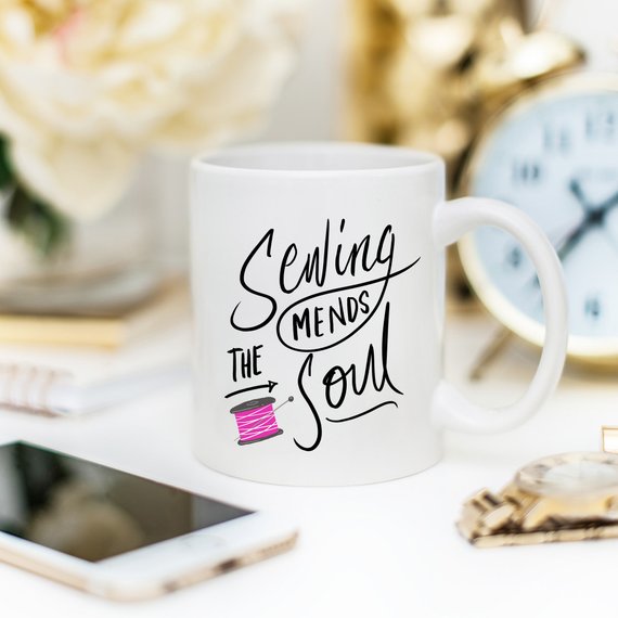 A ceramic coffee mug with the phrase 'Sewing Mends The Soul' printed in a fun design, perfect for sewing enthusiasts.