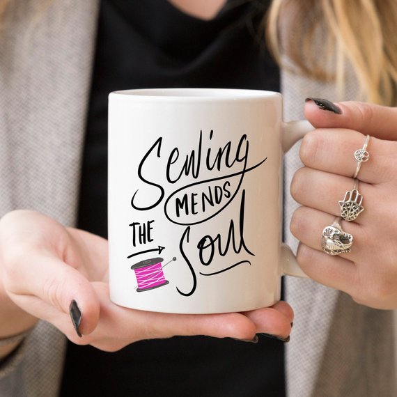 A ceramic coffee mug with the phrase 'Sewing Mends The Soul' printed in a fun design, perfect for sewing enthusiasts.