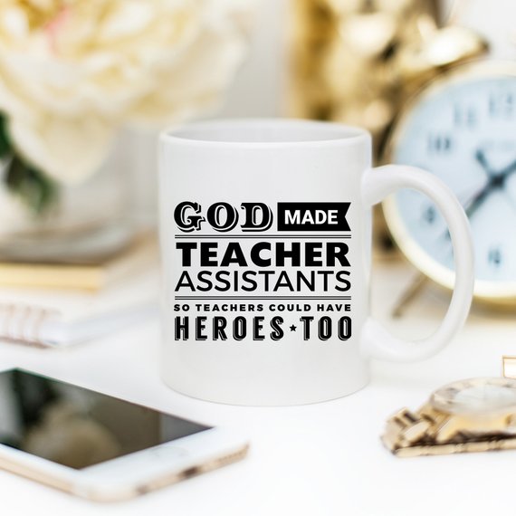 A white ceramic coffee mug with the text 'God Made Teacher Assistants So Teachers Could Have Heroes Too' printed in colorful letters, perfect for teacher assistants.