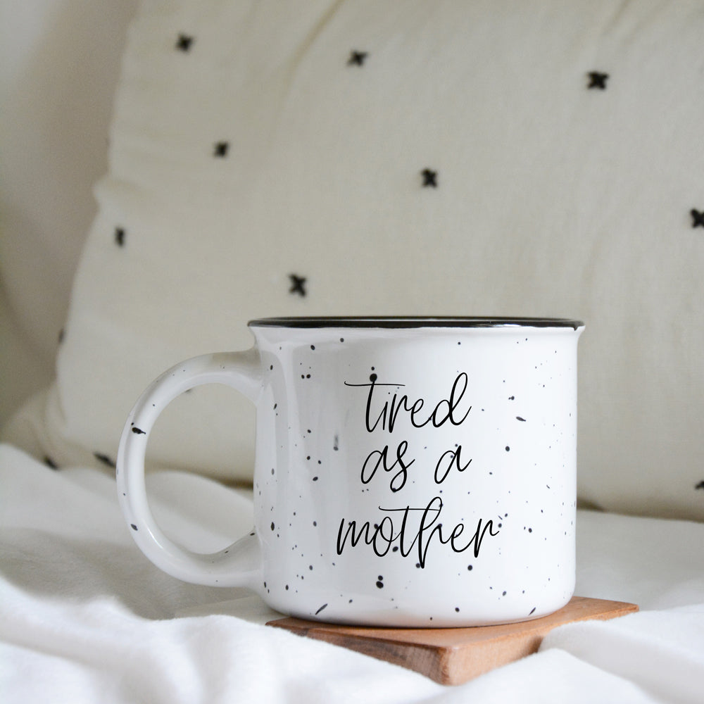 Tired as a Mother Campfire Mug with black rim and double-sided design, featuring white sesame glaze and bold black lettering.