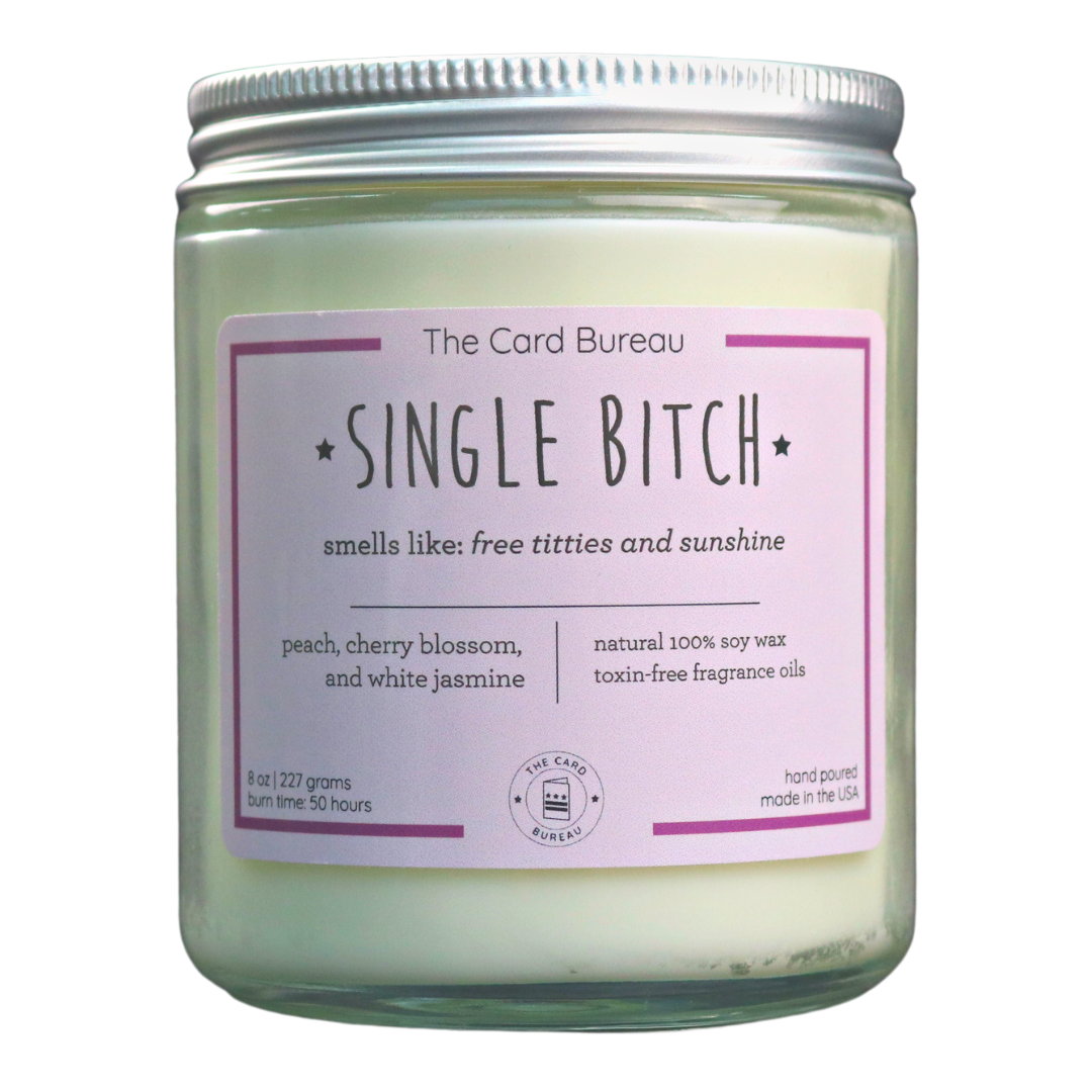 Single Bitch Candle in a glass jar with a tin lid, featuring a vibrant label and surrounded by decorative elements, showcasing its peach, cherry blossom, and jasmine scent.