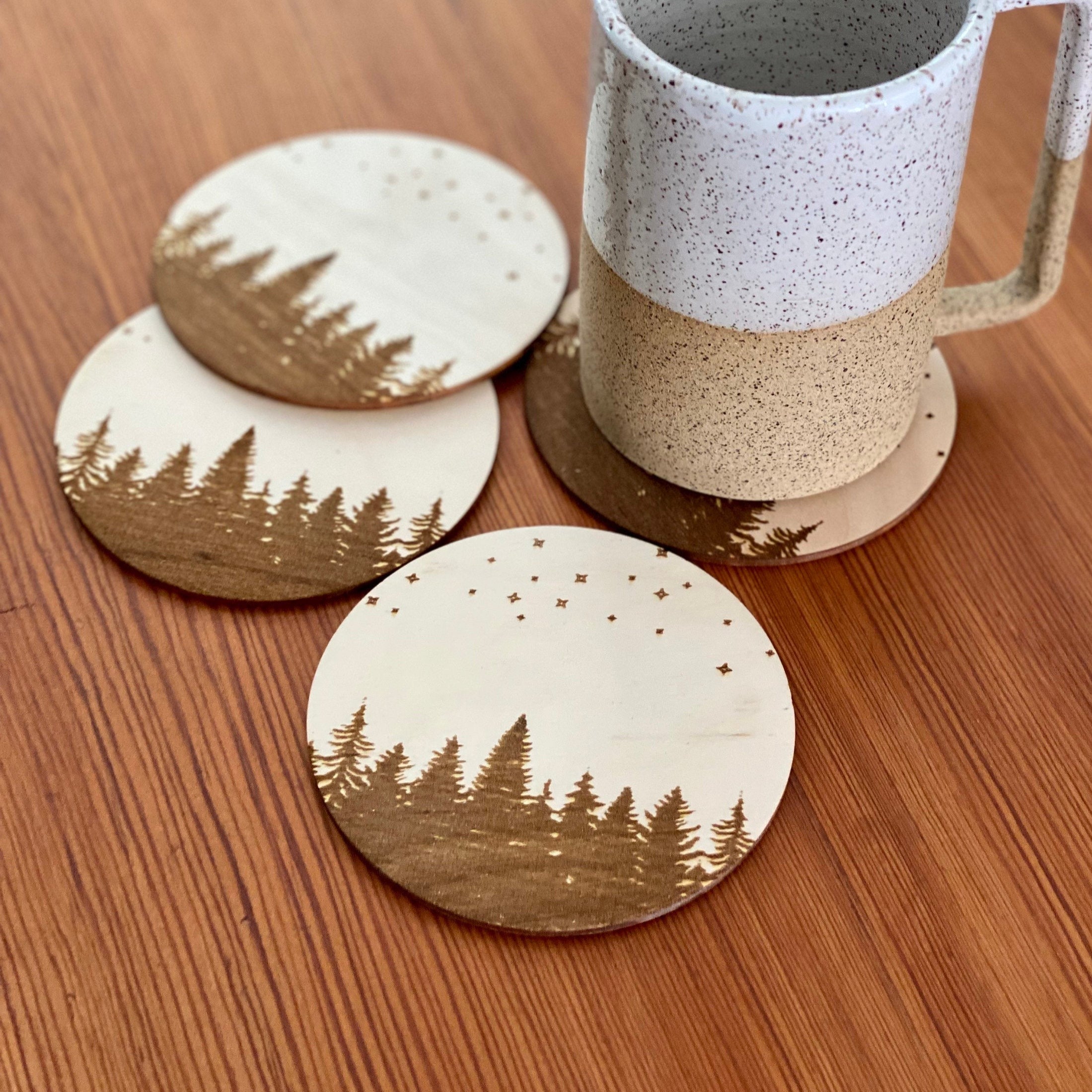 A set of four wooden coasters featuring intricate laser engravings of pine trees and a starry sky, showcasing their natural wood finish.