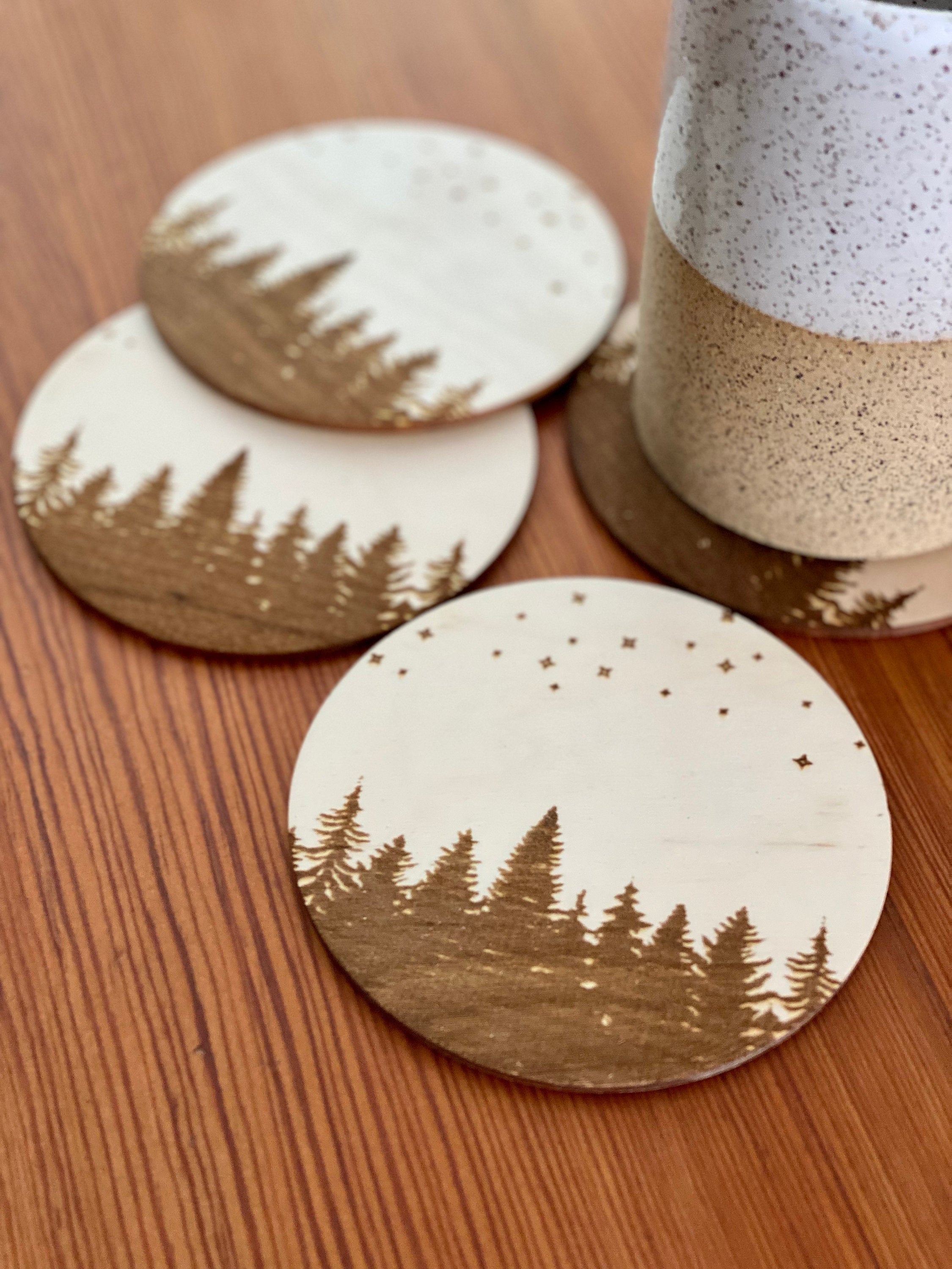 A set of four wooden coasters featuring intricate laser engravings of pine trees and a starry sky, showcasing their natural wood finish.