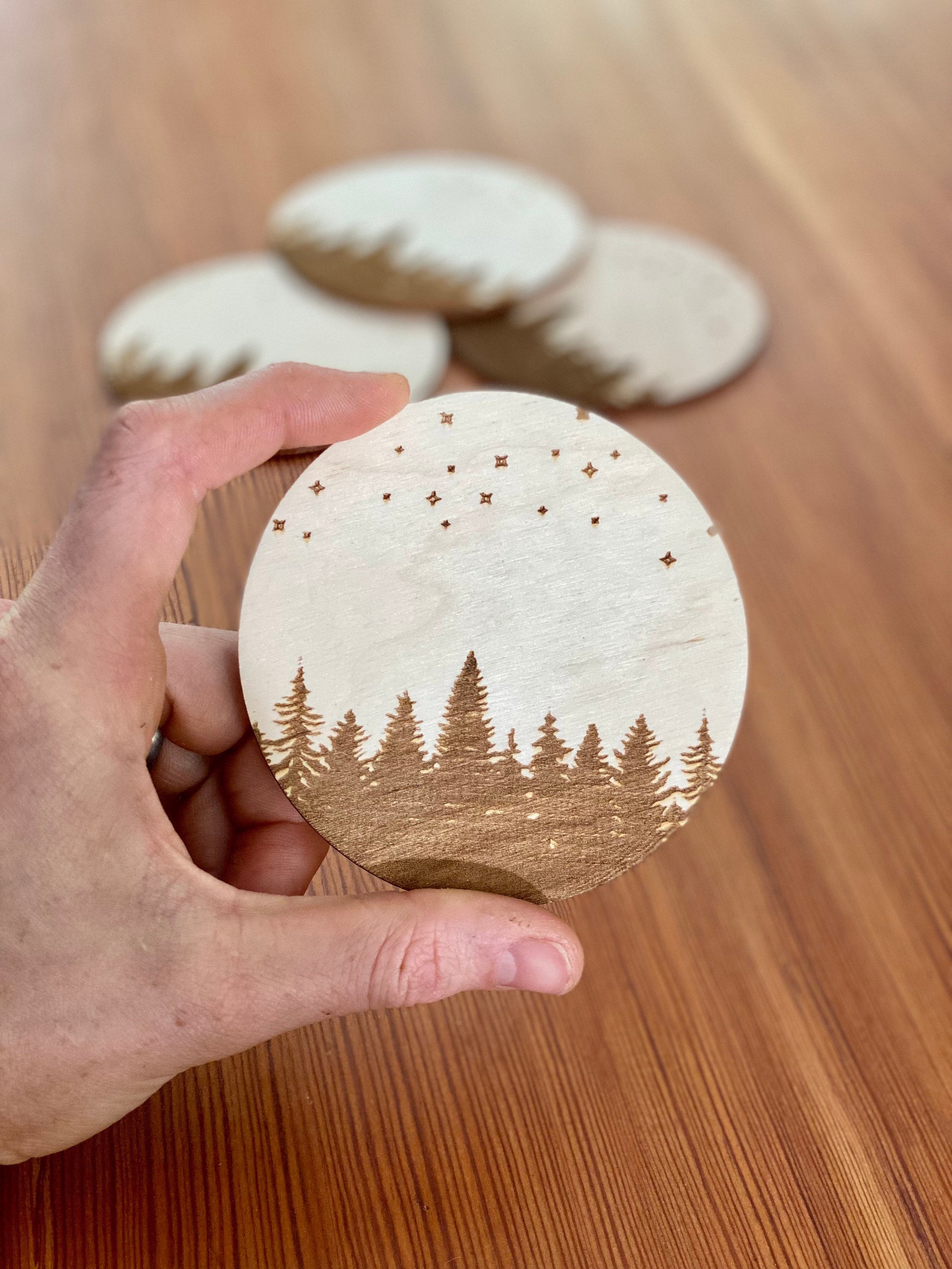 A set of four wooden coasters featuring intricate laser engravings of pine trees and a starry sky, showcasing their natural wood finish.