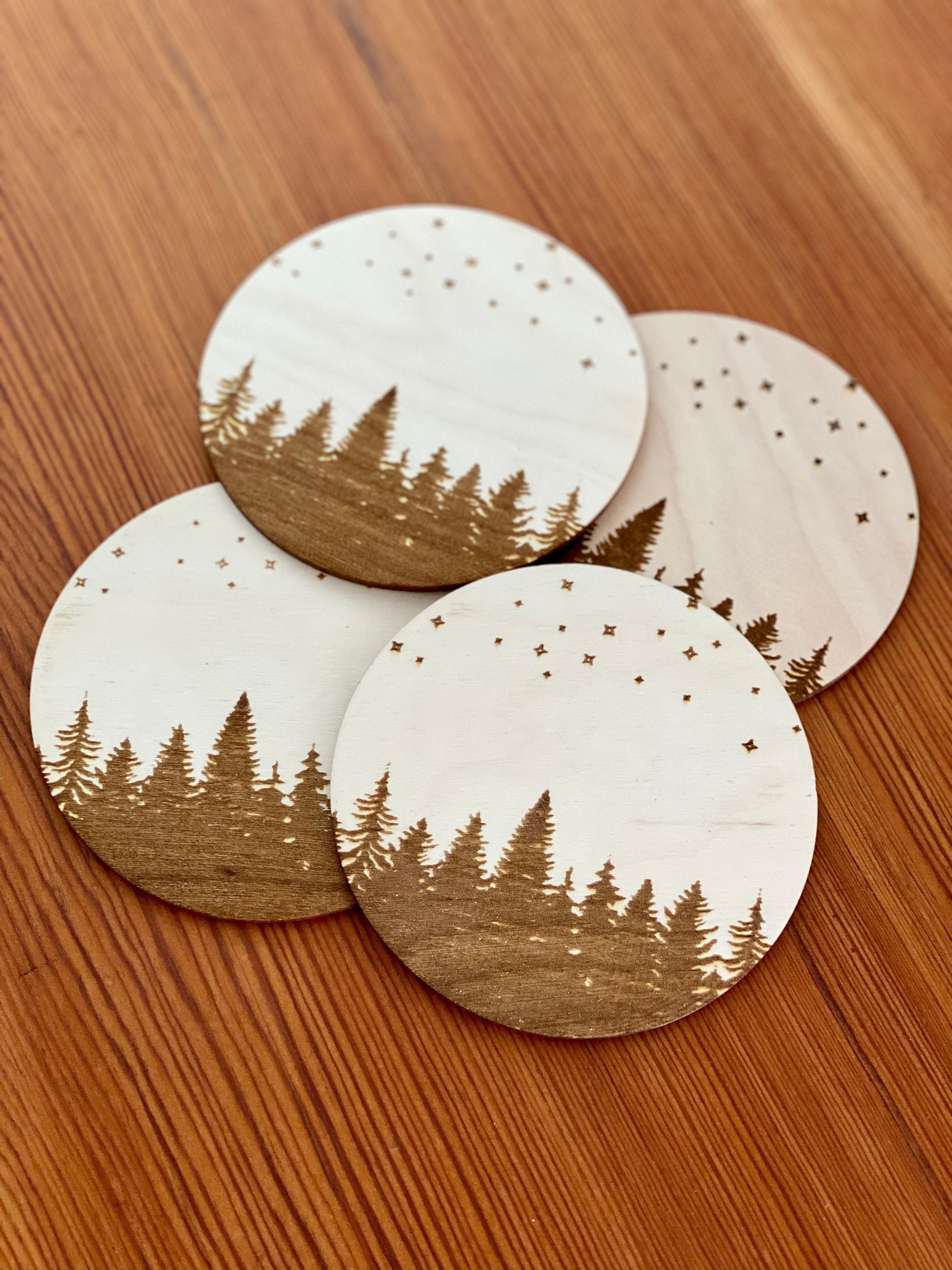 A set of four wooden coasters featuring intricate laser engravings of pine trees and a starry sky, showcasing their natural wood finish.