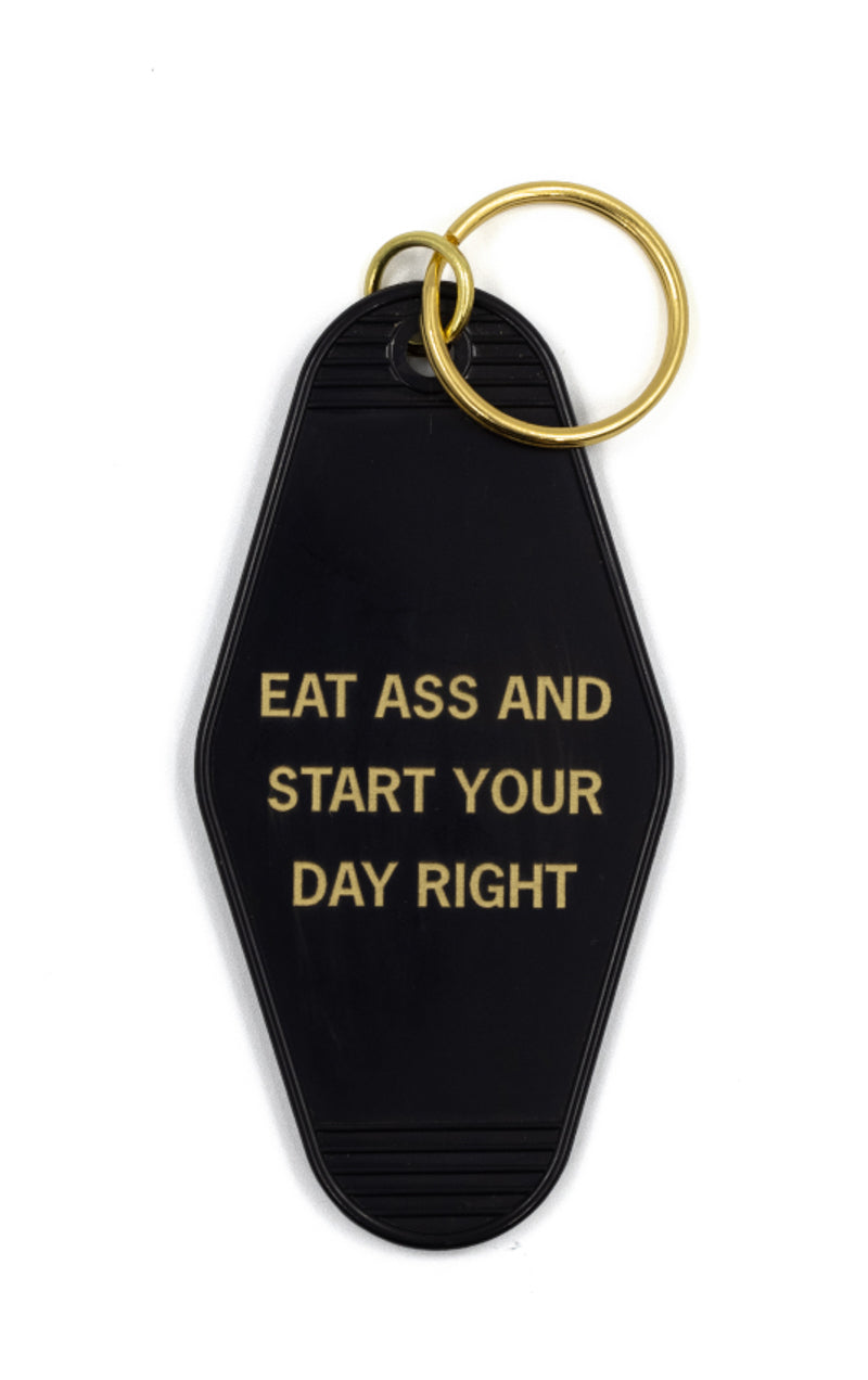 Black motel-style keychain with gold lettering that reads 'EAT ASS AND START YOUR DAY RIGHT', featuring a split ring for easy attachment.