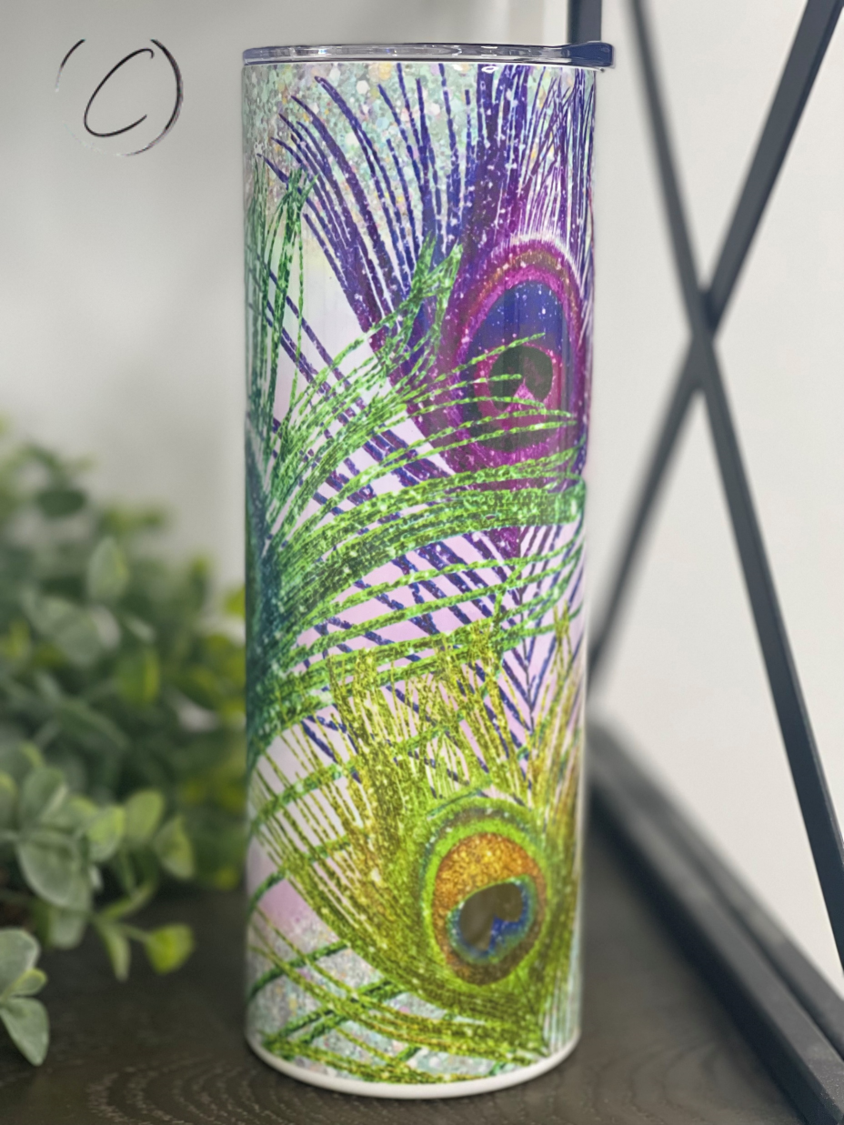 A 20oz skinny tumbler featuring a vibrant glitter peacock feather design, perfect for stylish hydration.