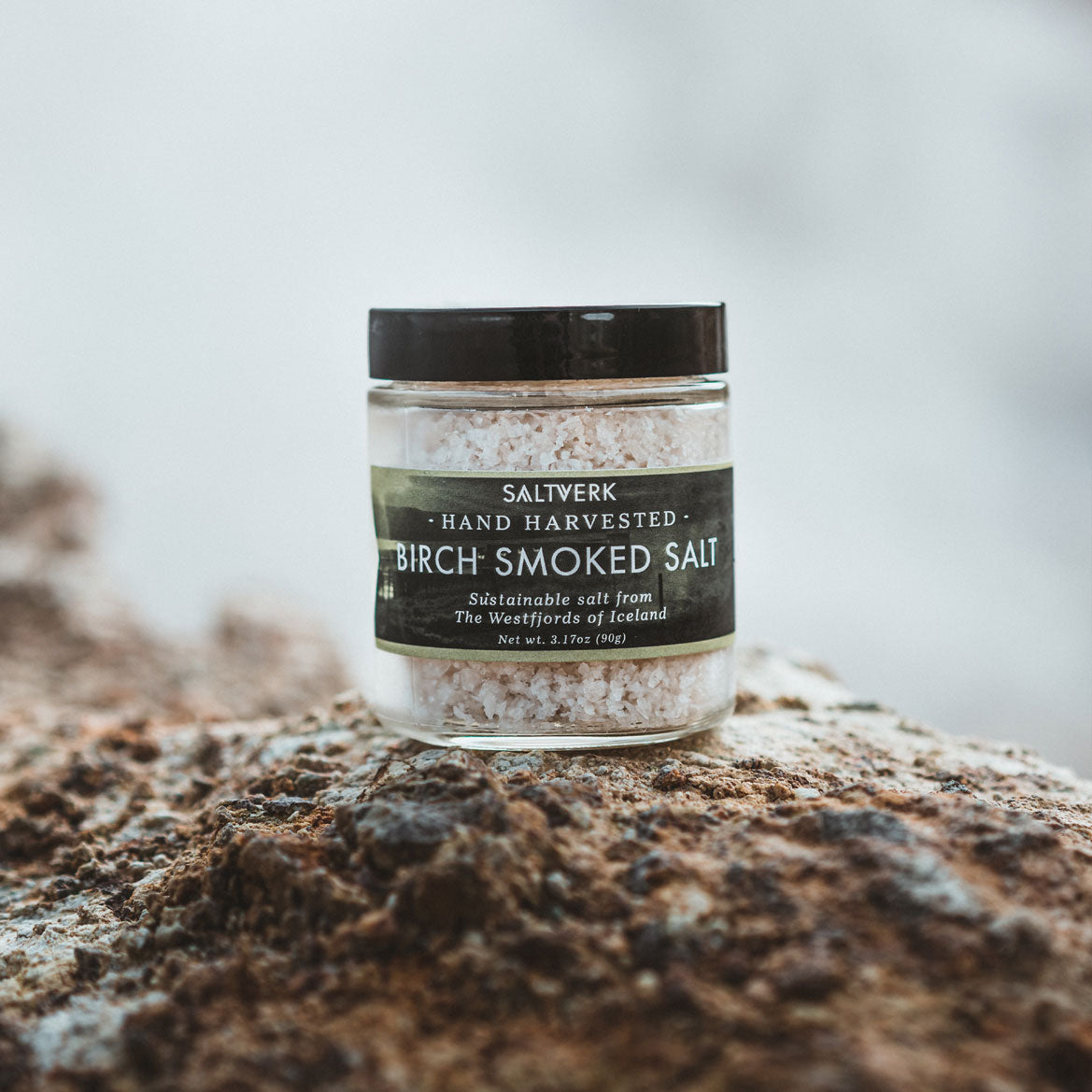 A jar of Birch Smoked Salt showcasing its coarse texture and rich color, surrounded by birch wood chips.