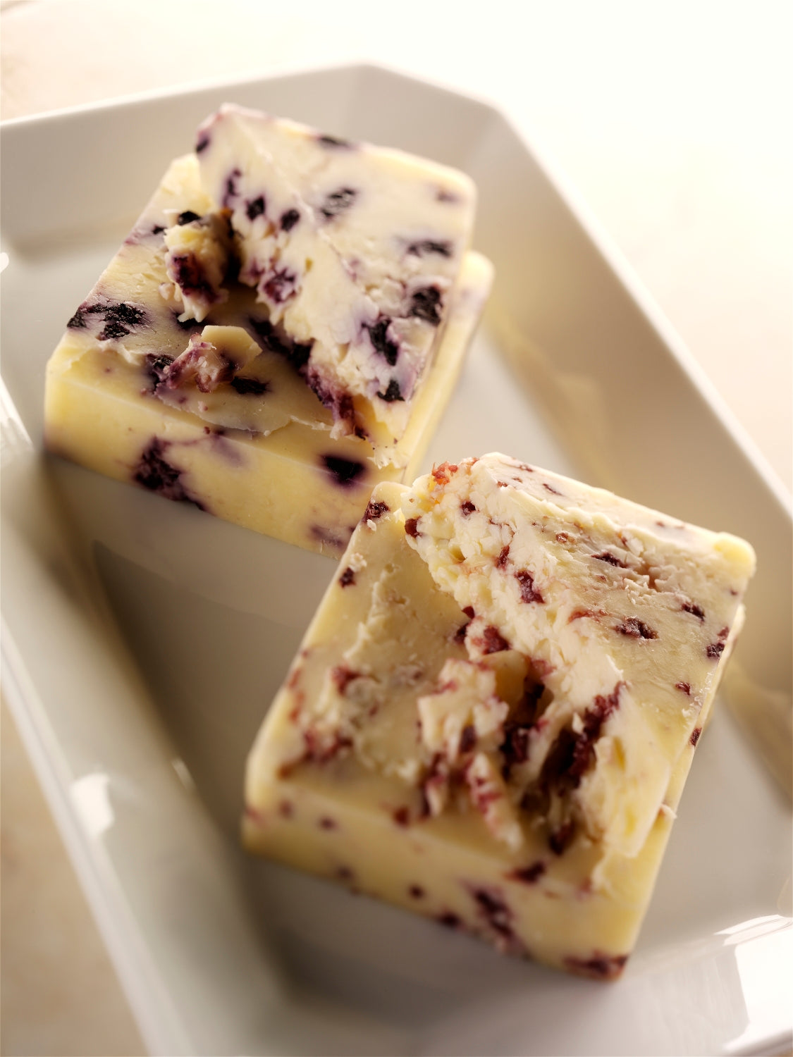 A wedge of Blueberry Infused Cheddar cheese showcasing its creamy texture and visible blueberry pieces, served on a wooden cheese board.