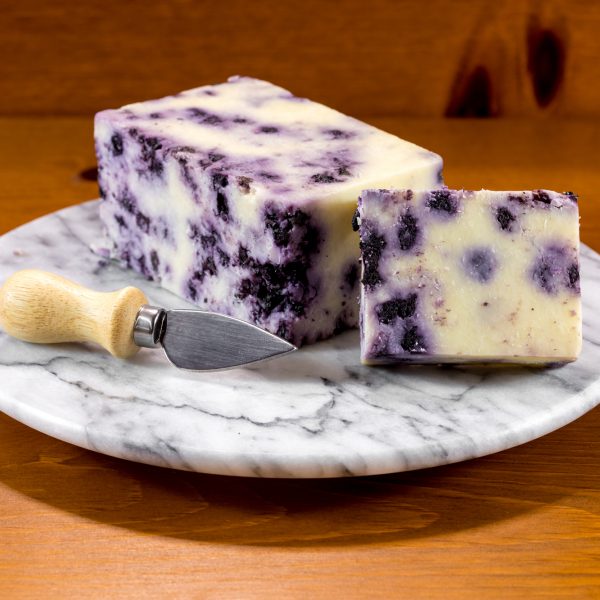 A wedge of Blueberry Infused Cheddar cheese showcasing its creamy texture and visible blueberry pieces, served on a wooden cheese board.