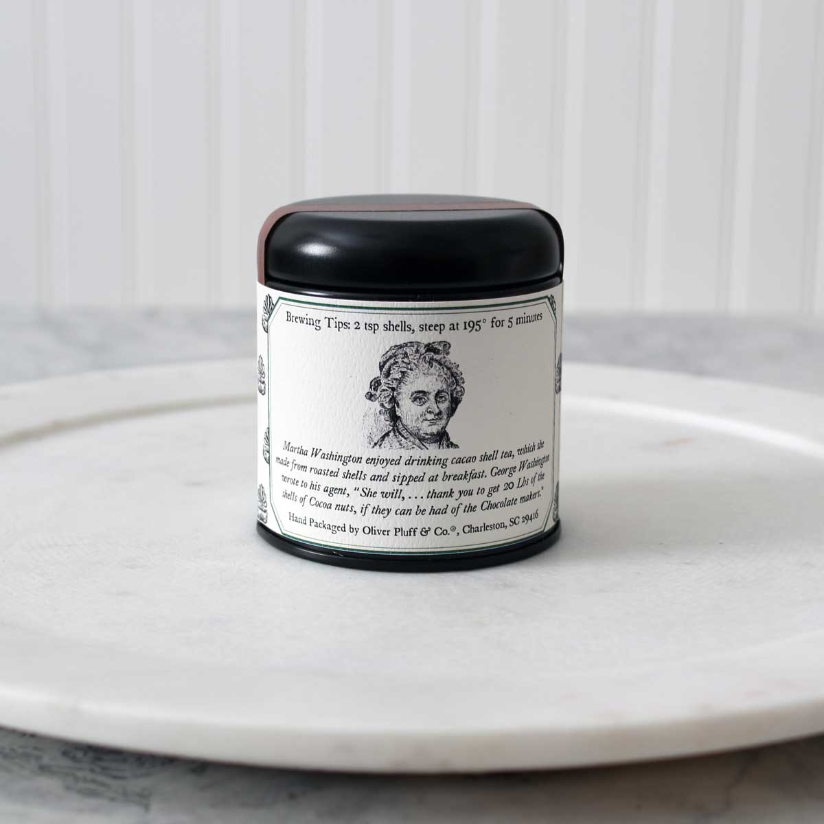 A tin of Martha Washington's Cacao Mint Tea containing loose roasted cacao shells blended with peppermint leaves, set against a rustic background.