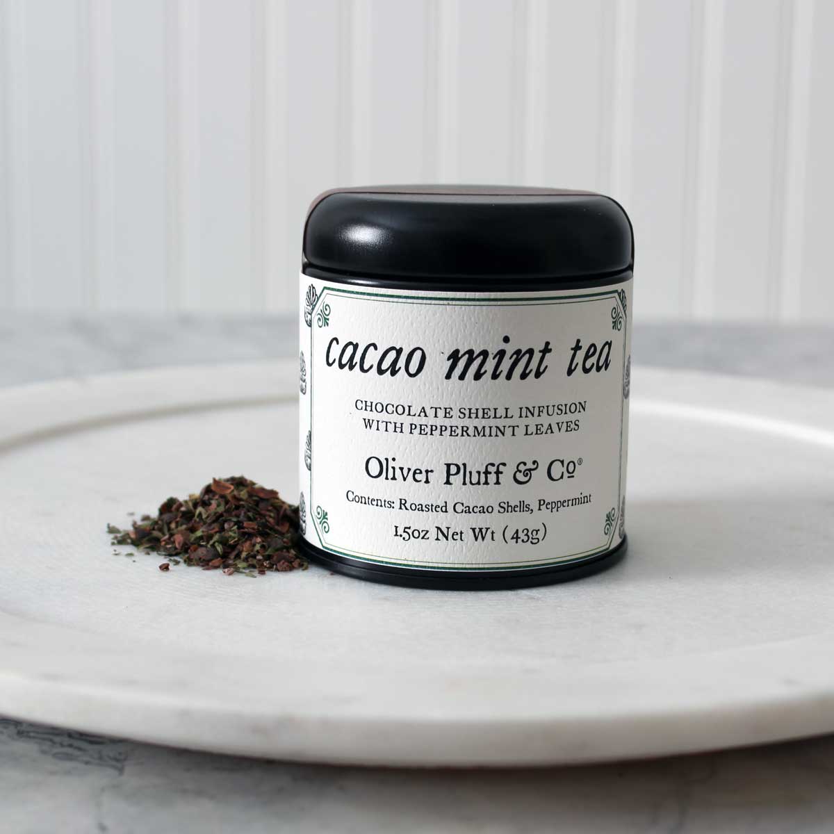 A tin of Martha Washington's Cacao Mint Tea containing loose roasted cacao shells blended with peppermint leaves, set against a rustic background.