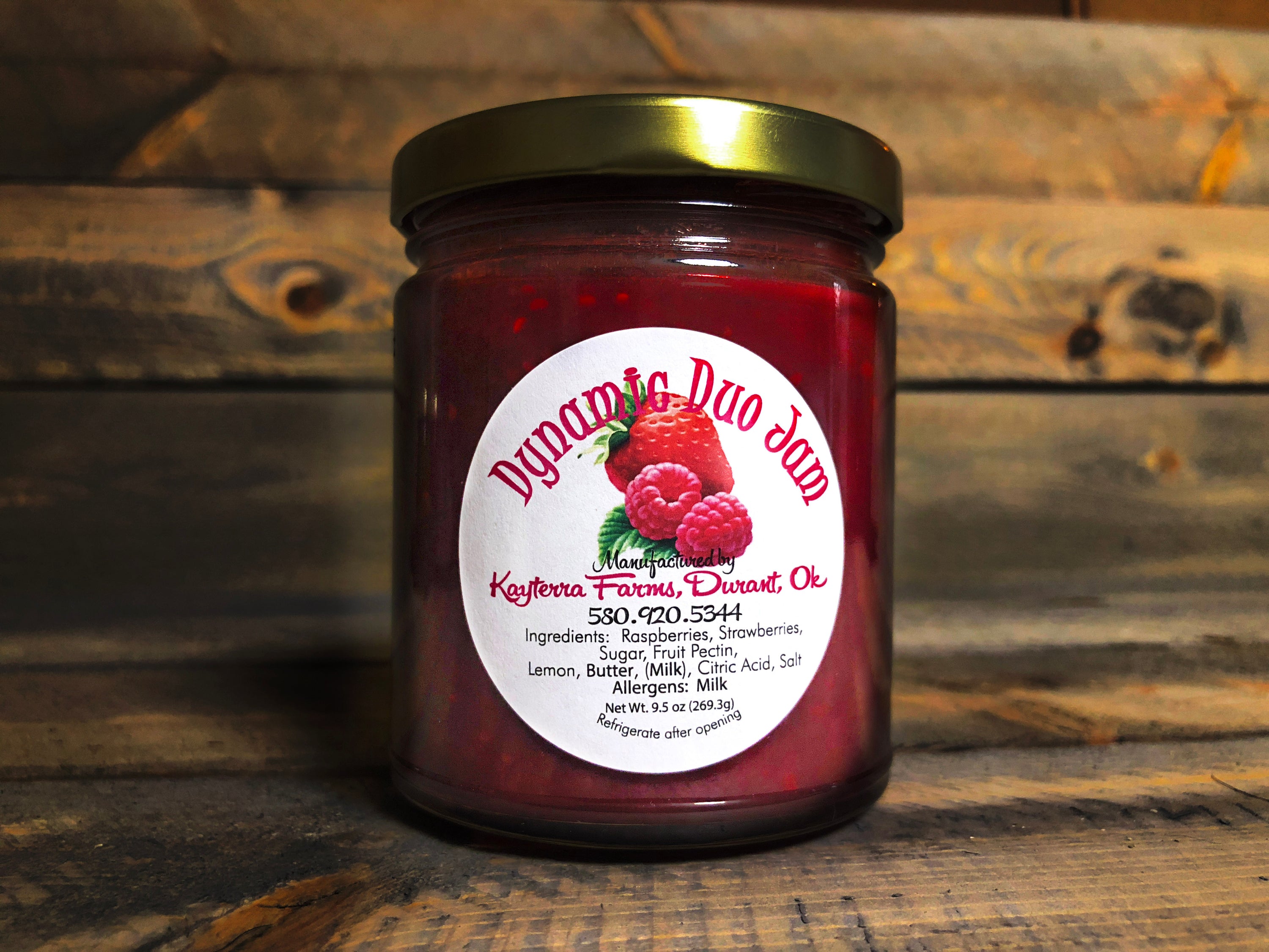 A jar of Dynamic Duo Strawberry/Raspberry jam, showcasing a vibrant red color with fresh strawberries and raspberries in the background.