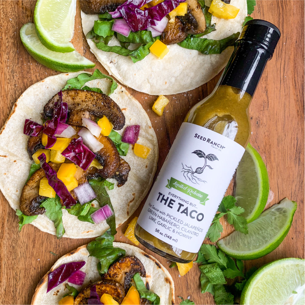 A bottle of Everything But The Taco seasoning with vibrant taco ingredients in the background, showcasing its flavorful essence.