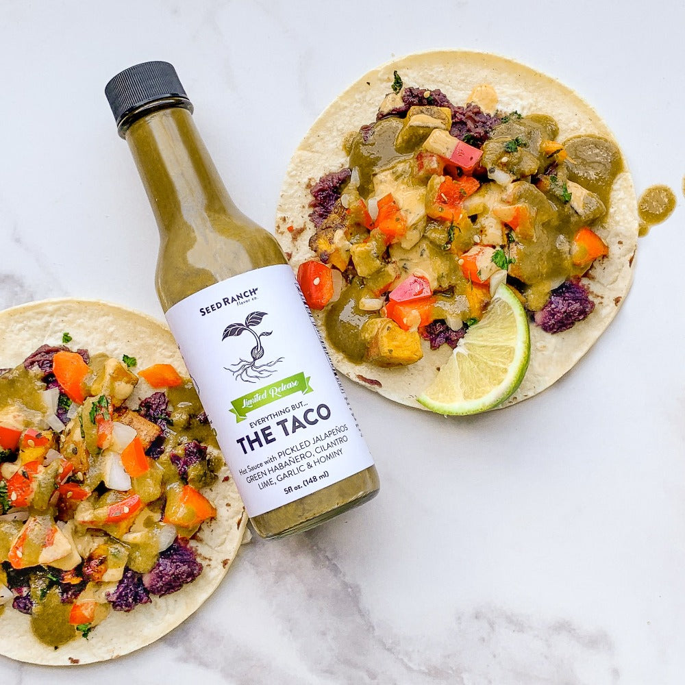 A bottle of Everything But The Taco seasoning with vibrant taco ingredients in the background, showcasing its flavorful essence.