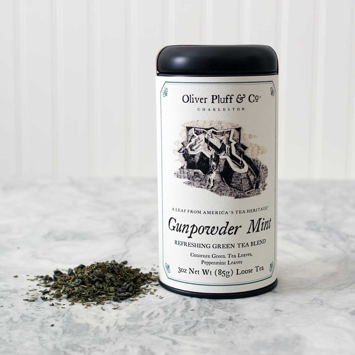 A matte black signature tea tin filled with Gunpowder Mint Loose Tea, showcasing tightly rolled green tea leaves and peppermint.