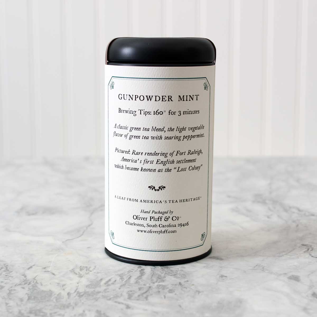 A matte black signature tea tin filled with Gunpowder Mint Loose Tea, showcasing tightly rolled green tea leaves and peppermint.