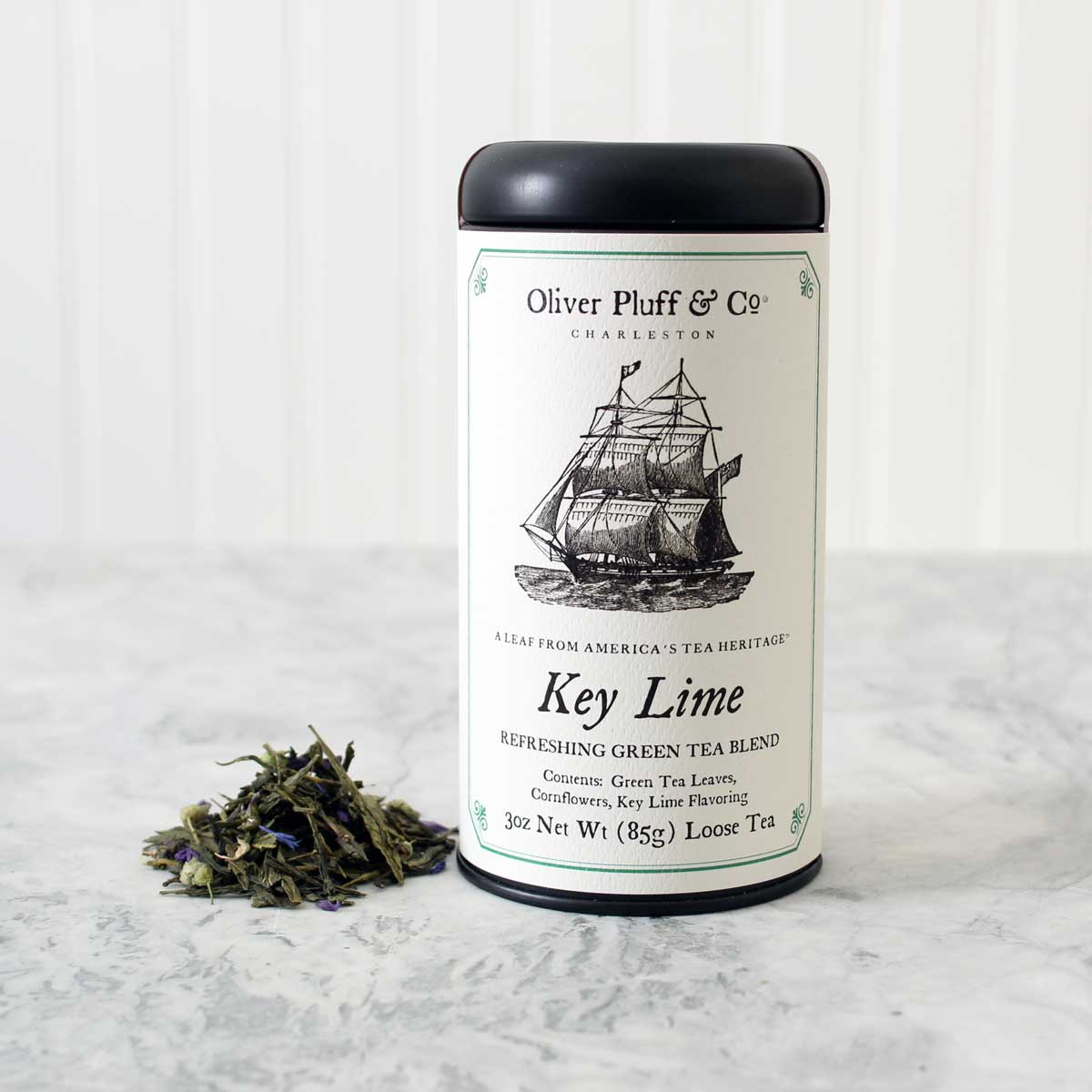 A stylish matte black tin containing Key Lime Loose Tea, showcasing large green tea leaves and blue mallow flowers, perfect for tea enthusiasts.