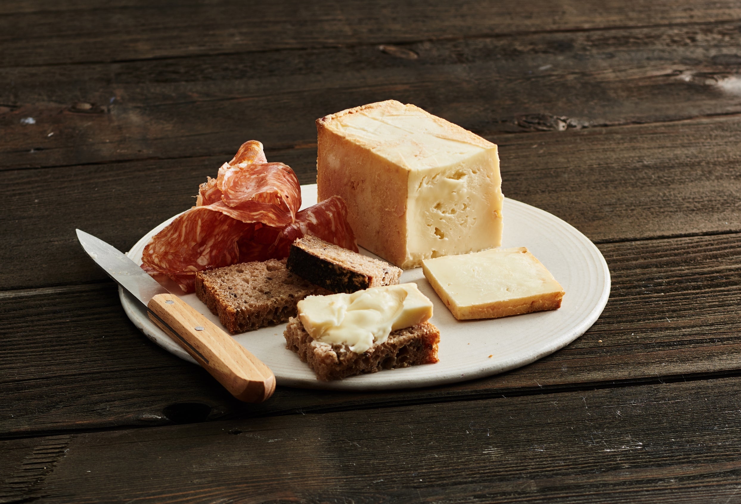 A round wheel of Limburger cheese with a creamy texture and strong aroma, displayed on a wooden cheese board alongside slices of bread and a glass of Zinfandel wine.