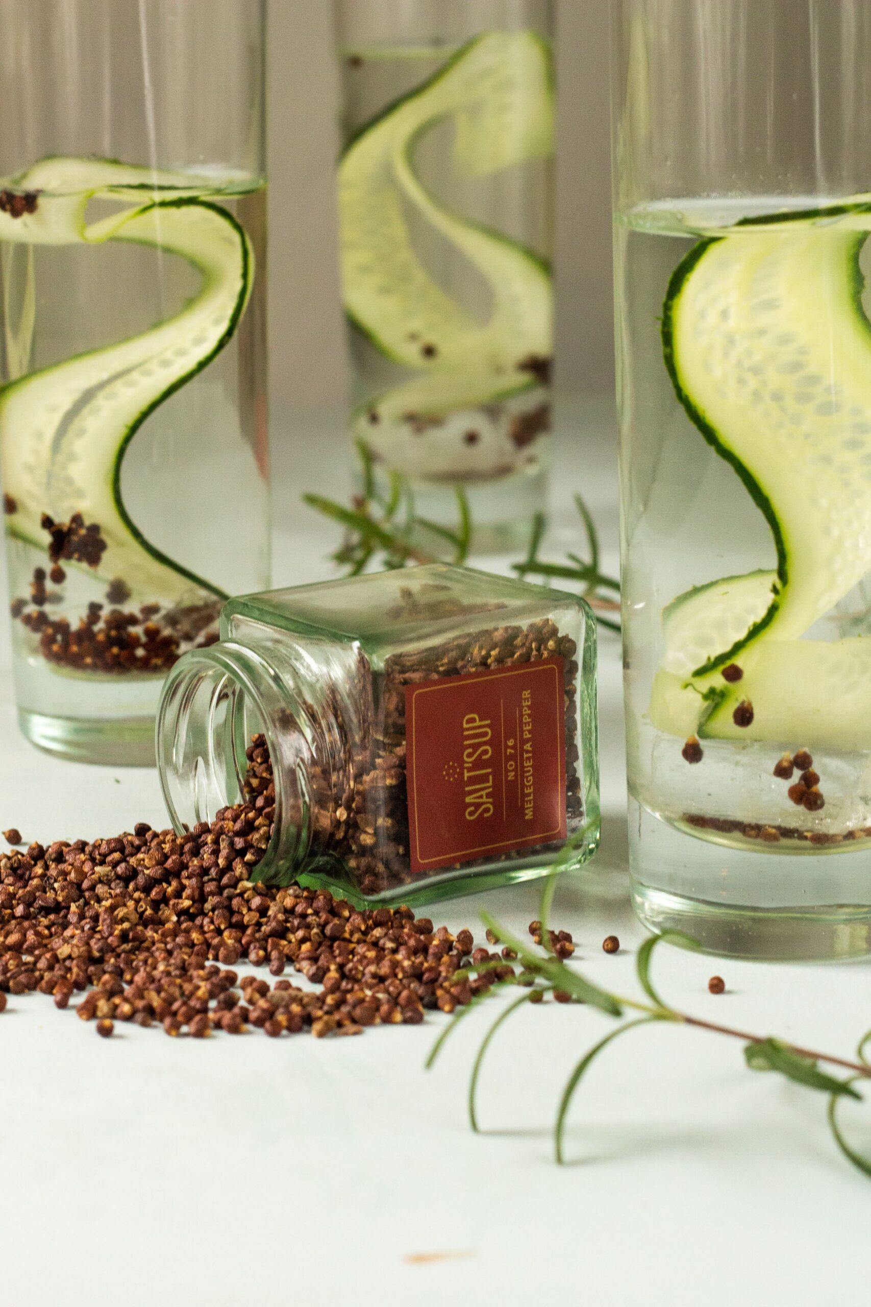 A glass jar filled with Melegueta peppercorns, showcasing their unique shape and color, with a background of spices and herbs.