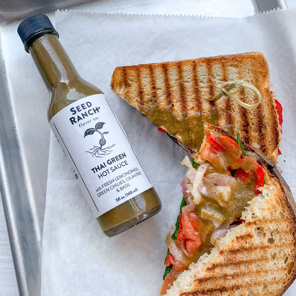 A bottle of Thai Green Hot Sauce featuring a vibrant green color, with fresh ingredients like lemongrass and lime in the background.