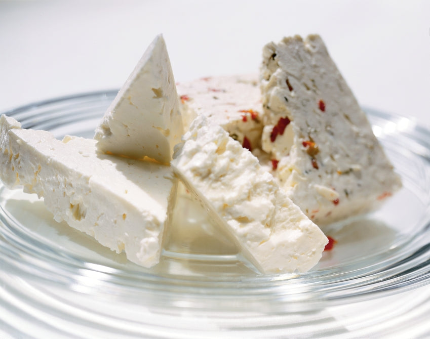 A block of Tomato Basil Feta cheese with fresh tomatoes and basil leaves, showcasing its vibrant colors and textures.