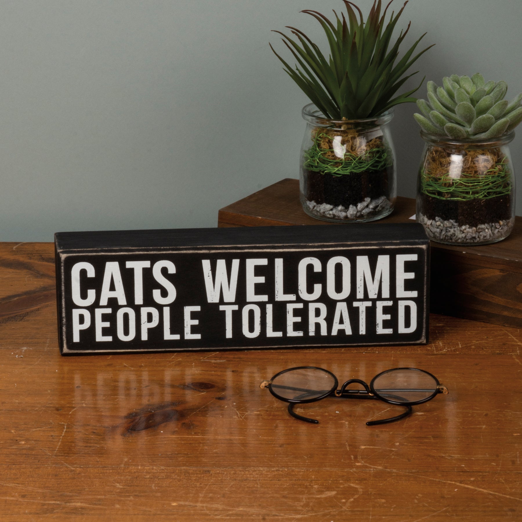 A rustic black and white wooden box sign that reads 'Cats Welcome - People Tolerated', featuring a distressed finish.