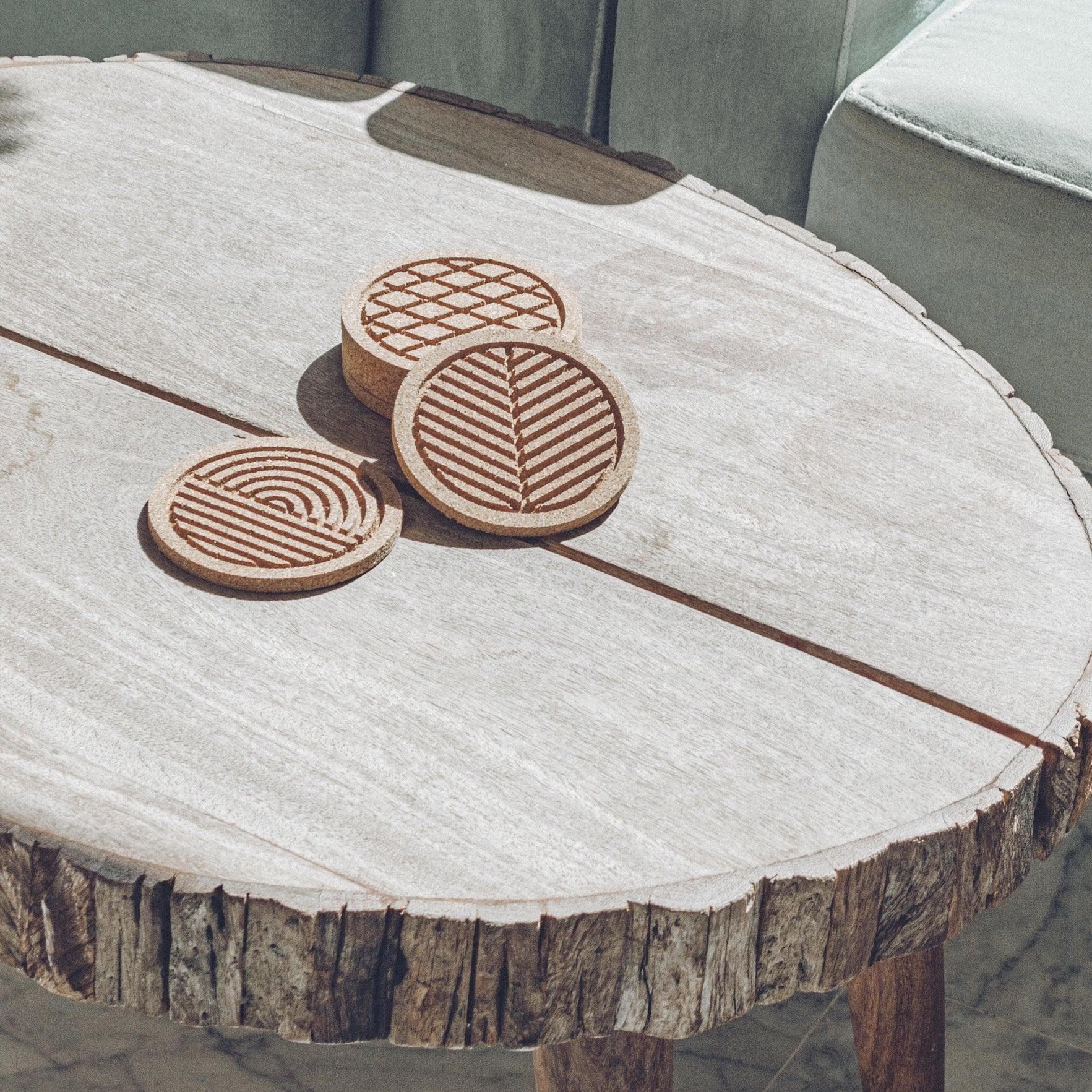 A set of six round cork coasters with nature-inspired designs including waves, leaves, and mountains, elegantly displayed on a wooden table.