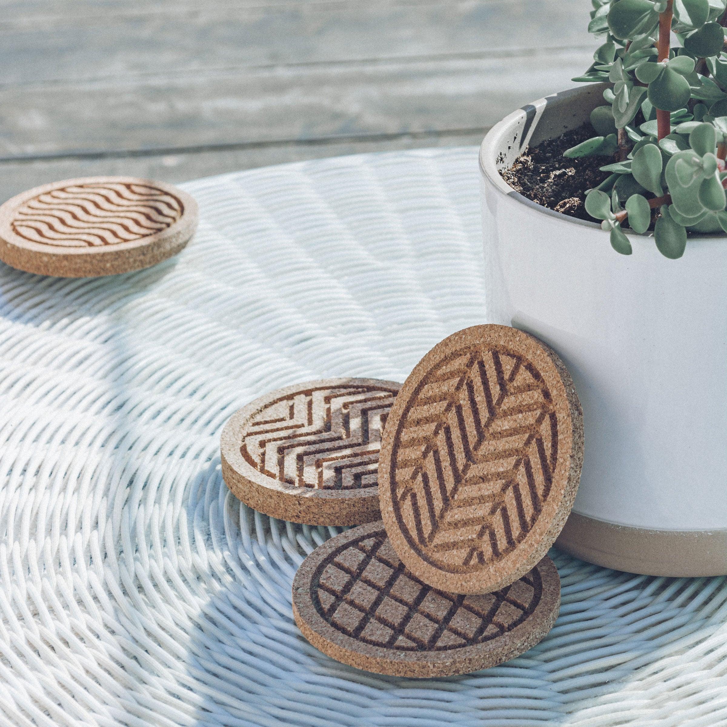 A set of six round cork coasters with nature-inspired designs including waves, leaves, and mountains, elegantly displayed on a wooden table.