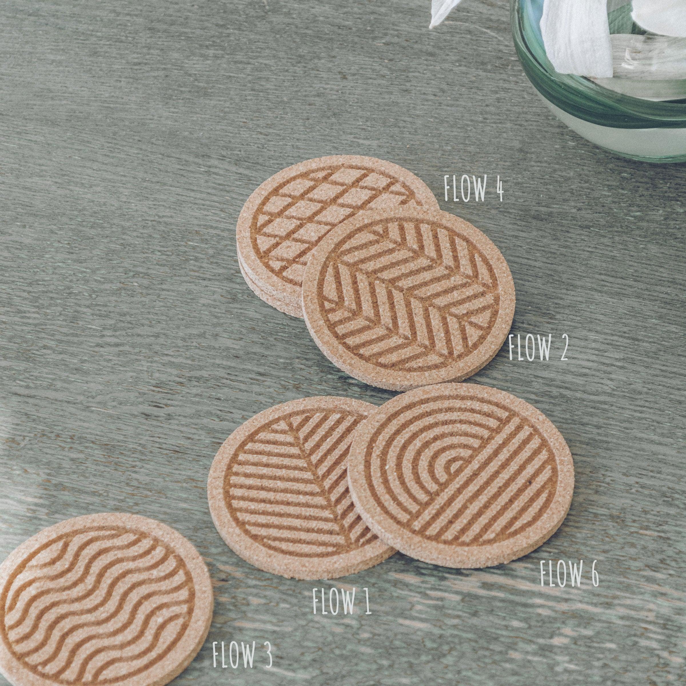 A set of six round cork coasters with nature-inspired designs including waves, leaves, and mountains, elegantly displayed on a wooden table.