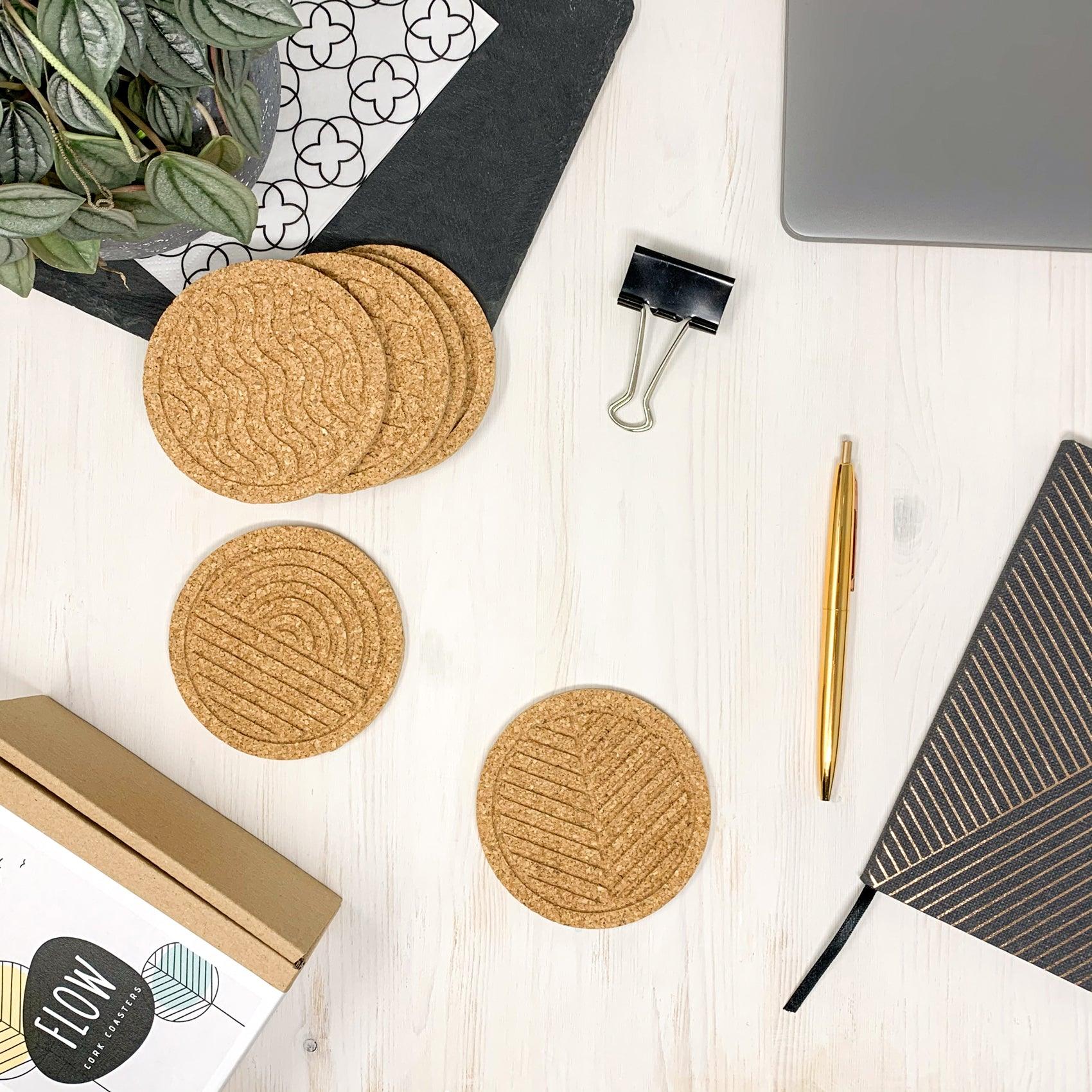 A set of six round cork coasters with nature-inspired designs including waves, leaves, and mountains, elegantly displayed on a wooden table.