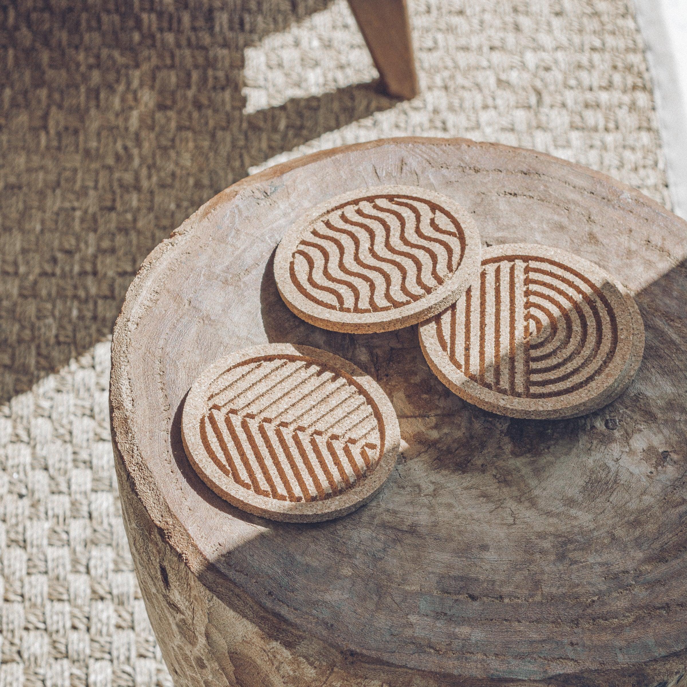 A set of six round cork coasters with nature-inspired designs including waves, leaves, and mountains, elegantly displayed on a wooden table.