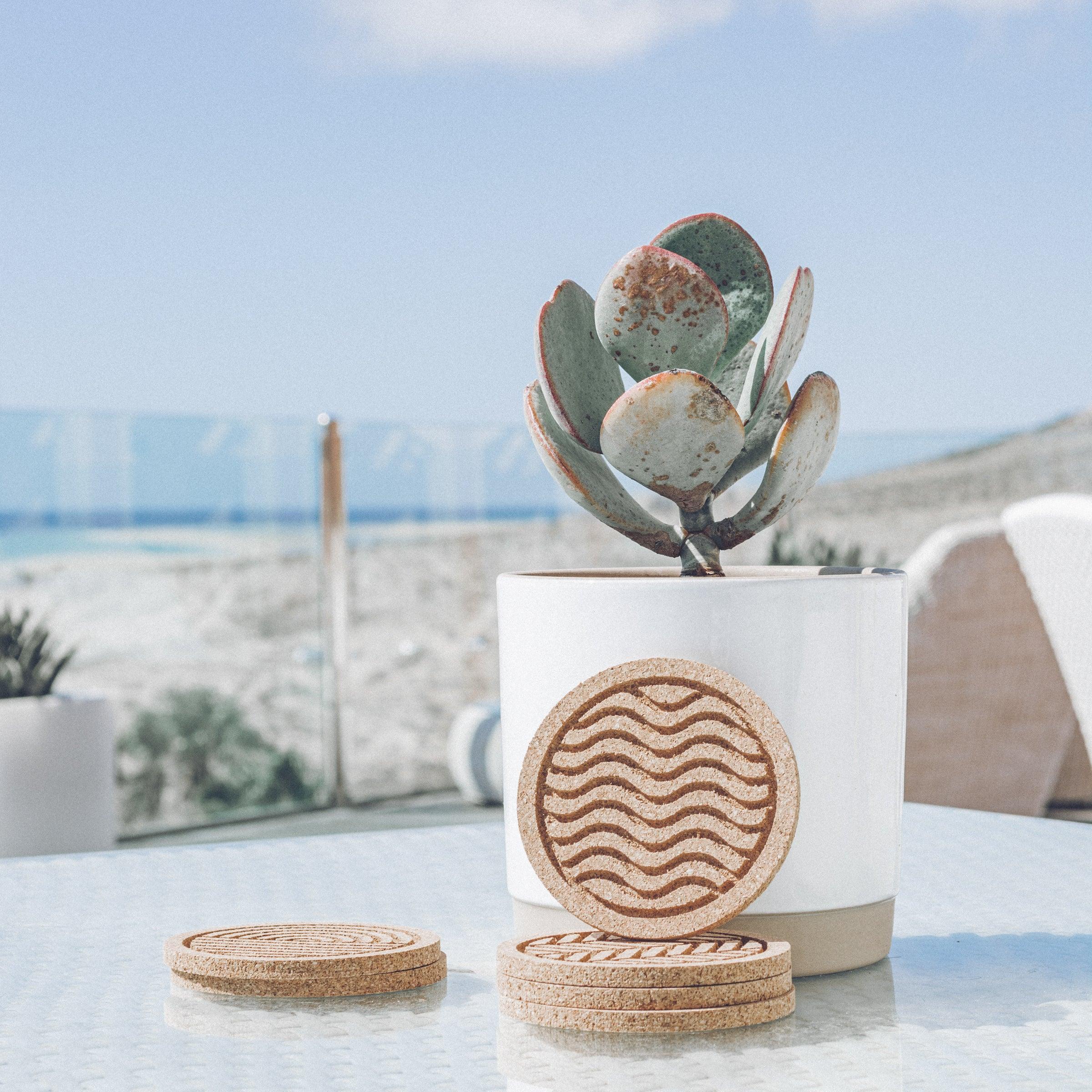 A set of six round cork coasters with nature-inspired designs including waves, leaves, and mountains, elegantly displayed on a wooden table.