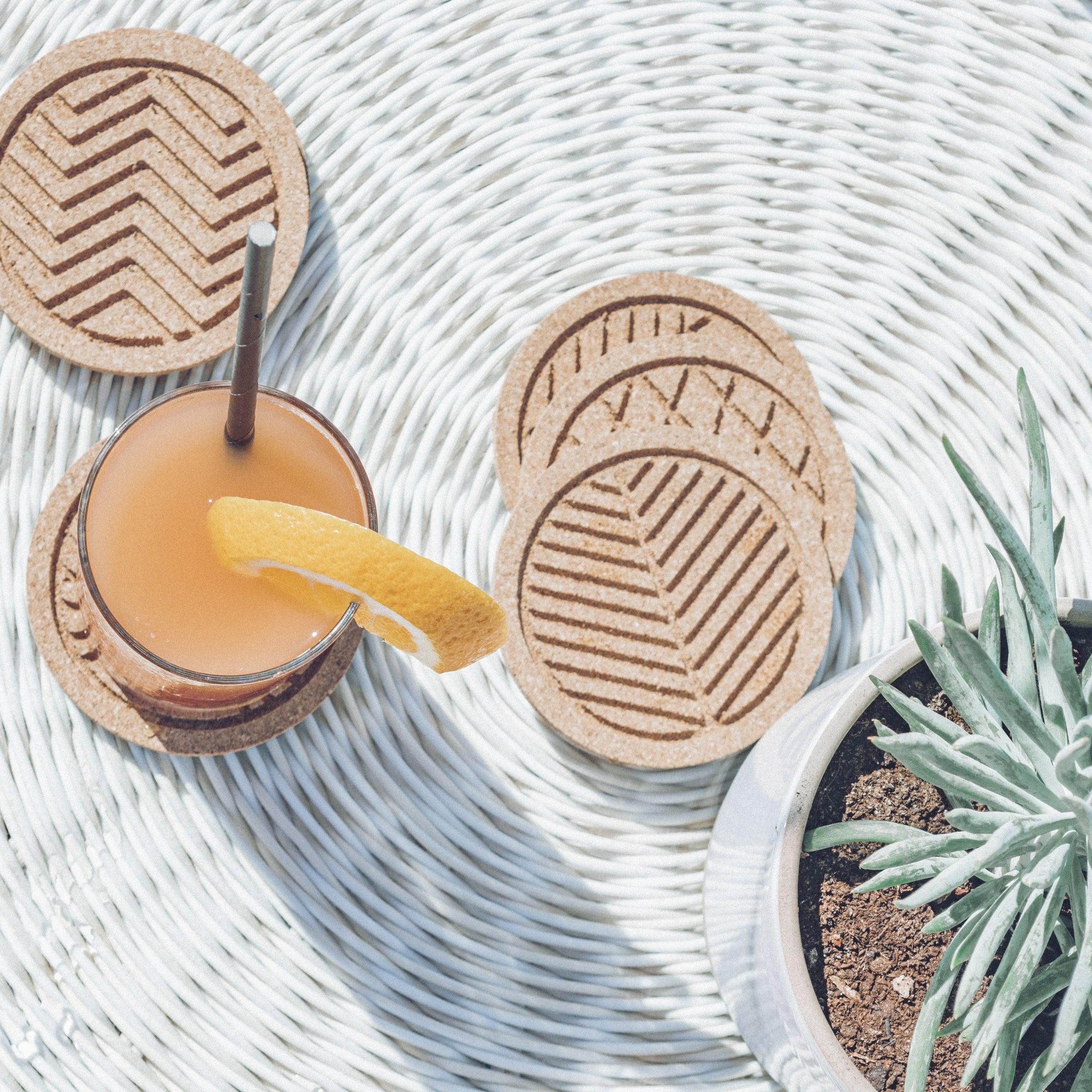 A set of six round cork coasters with nature-inspired designs including waves, leaves, and mountains, elegantly displayed on a wooden table.