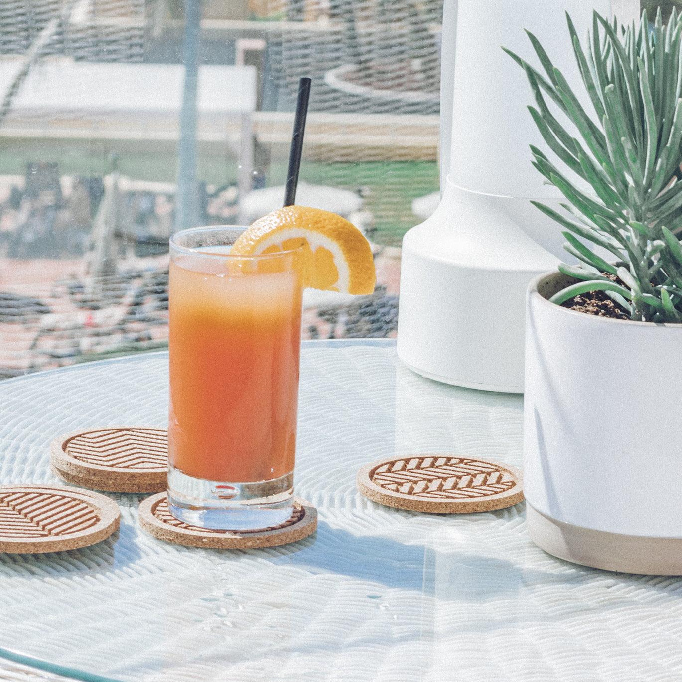 A set of six round cork coasters with nature-inspired designs including waves, leaves, and mountains, elegantly displayed on a wooden table.