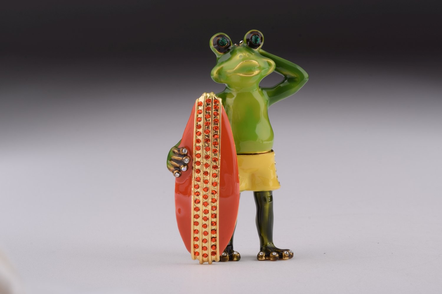 A beautifully crafted trinket box shaped like a frog holding a surfboard, adorned with Austrian crystals and plated with gold.