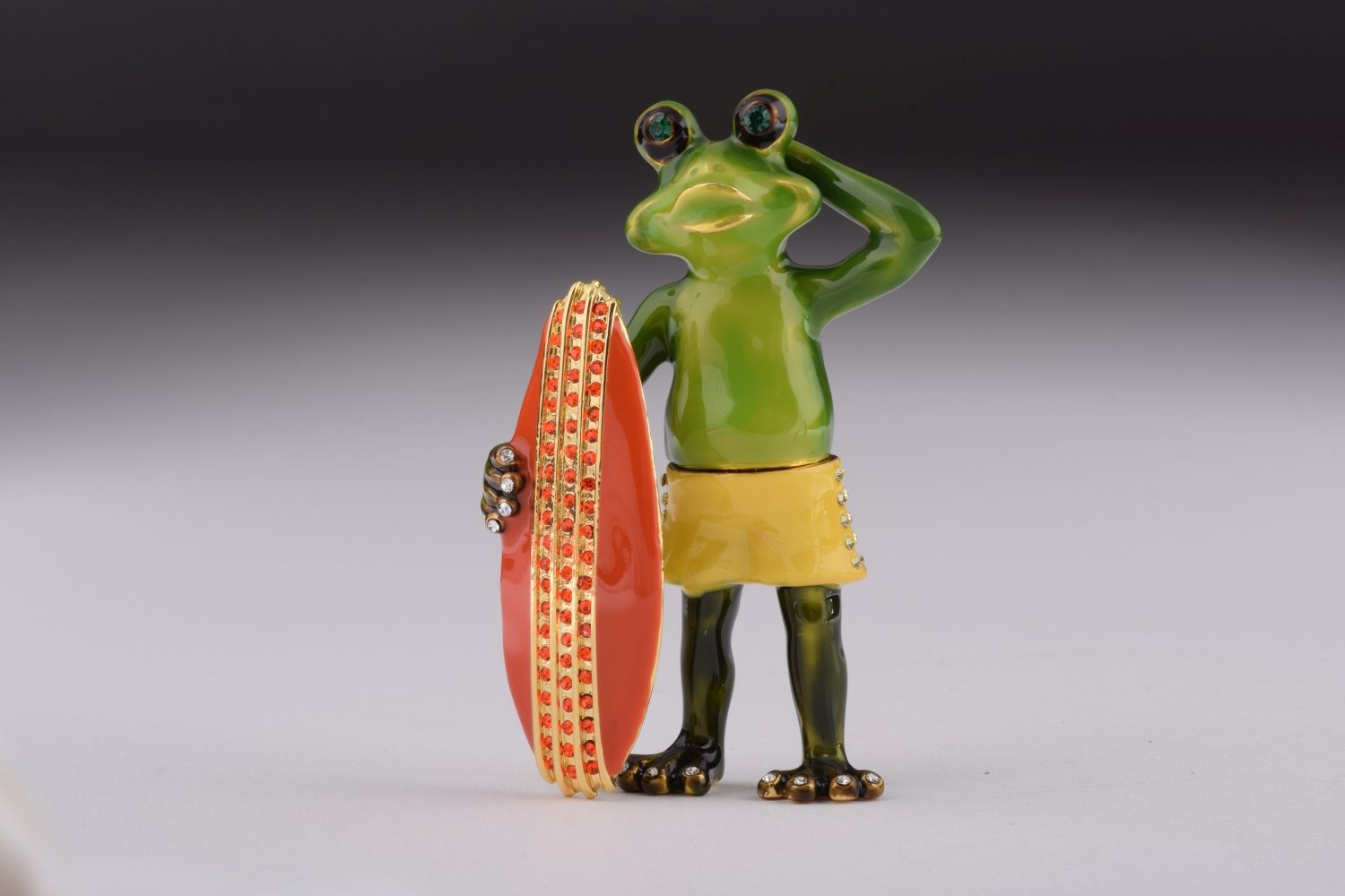 A beautifully crafted trinket box shaped like a frog holding a surfboard, adorned with Austrian crystals and plated with gold.