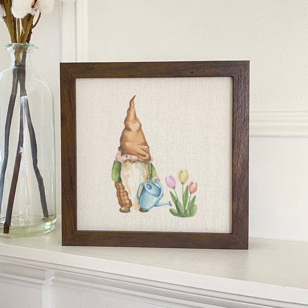 A charming framed sign featuring a garden gnome surrounded by tulips, with a natural wood frame and eco-friendly printing.