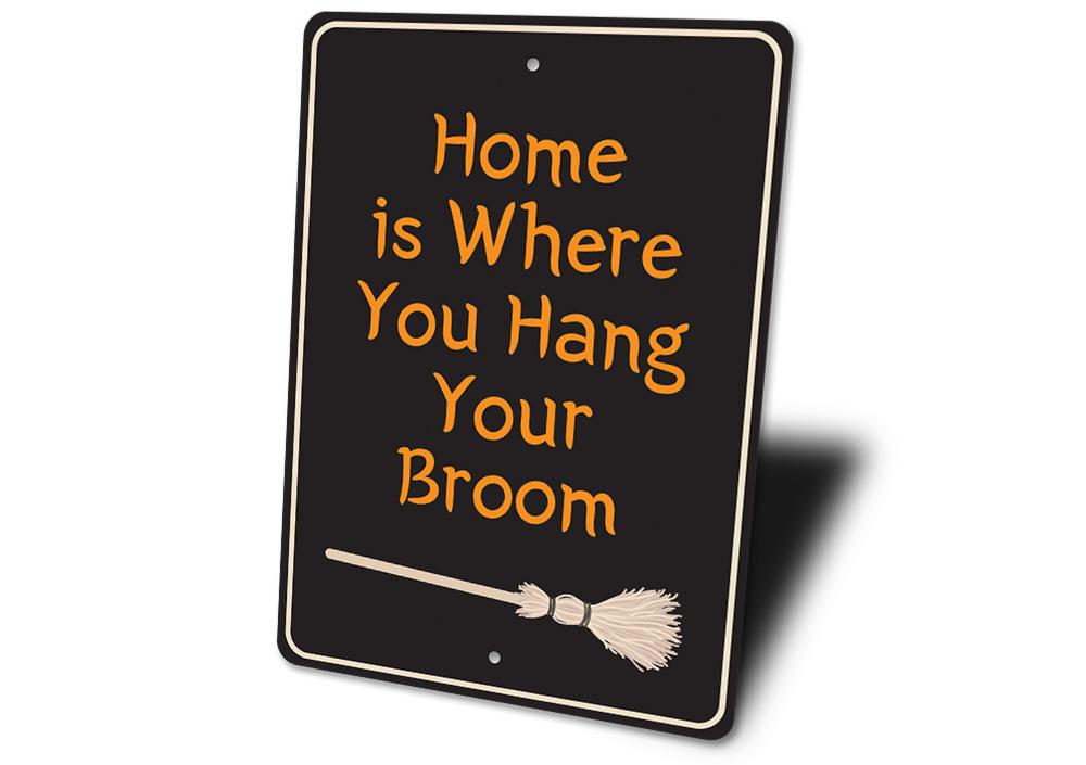 A decorative metal sign reading 'Home is Where You Hang Your Broom', featuring spooky Halloween-themed design elements.