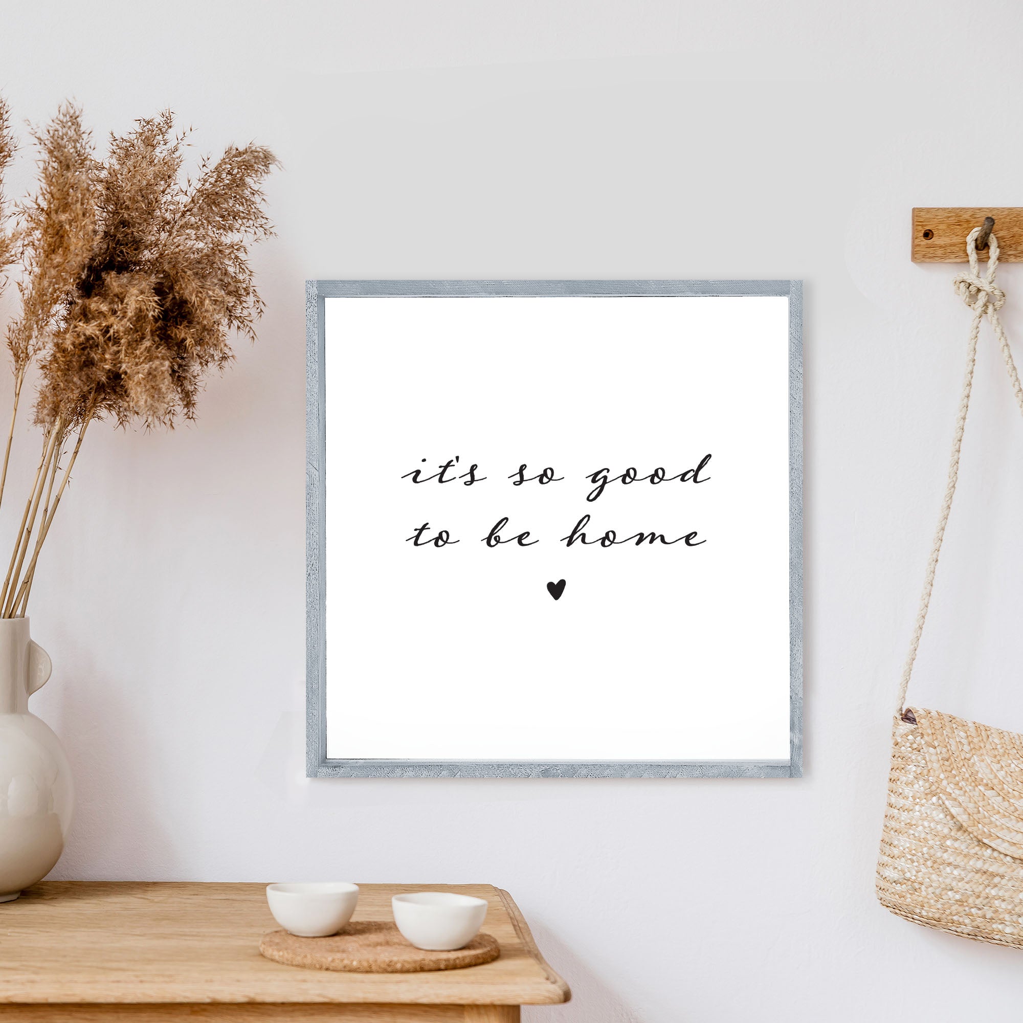 A charming 13x13 inch wood sign with the phrase 'It's So Good To Be Home' in black text on a white background, framed in stained pine wood.