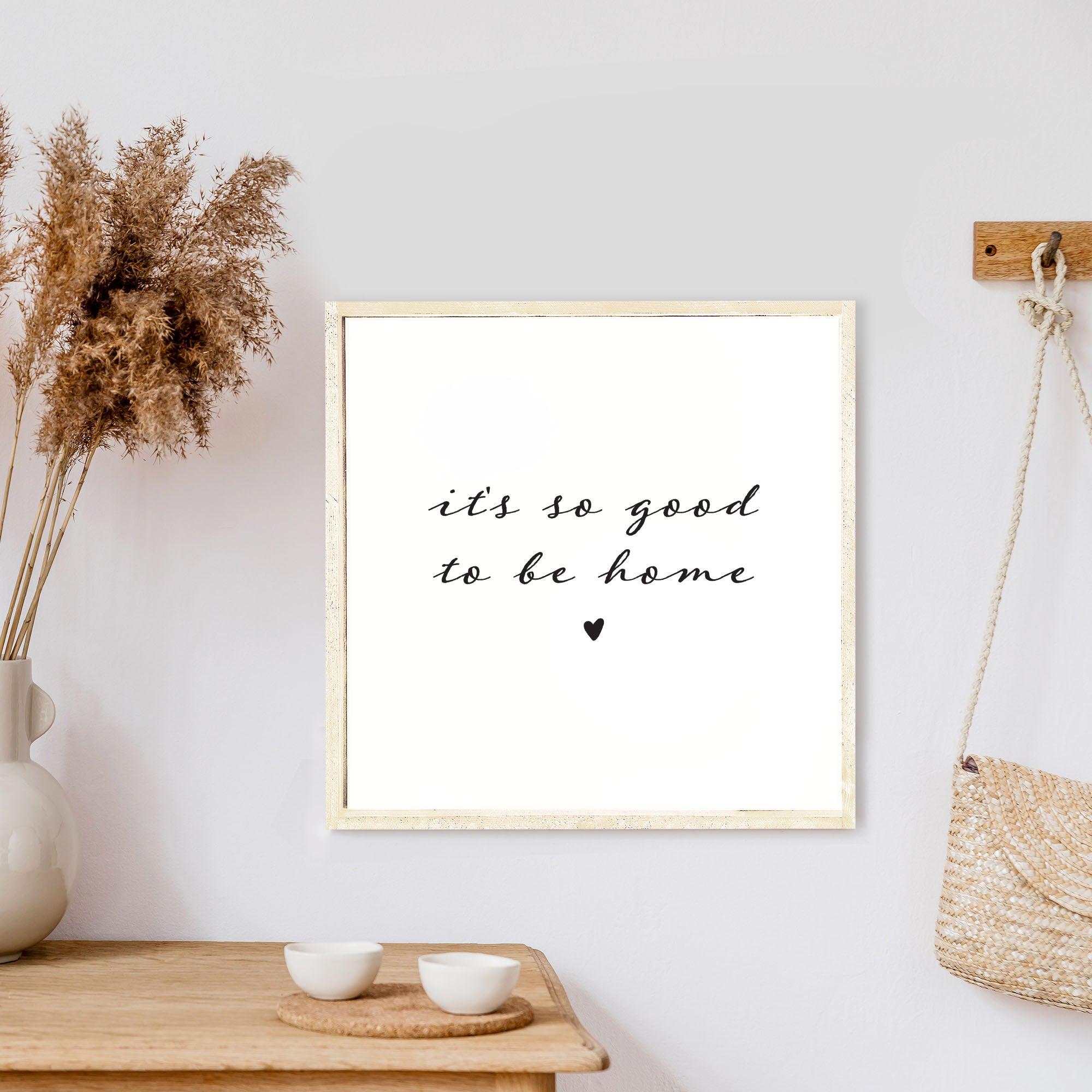 A charming 13x13 inch wood sign with the phrase 'It's So Good To Be Home' in black text on a white background, framed in stained pine wood.