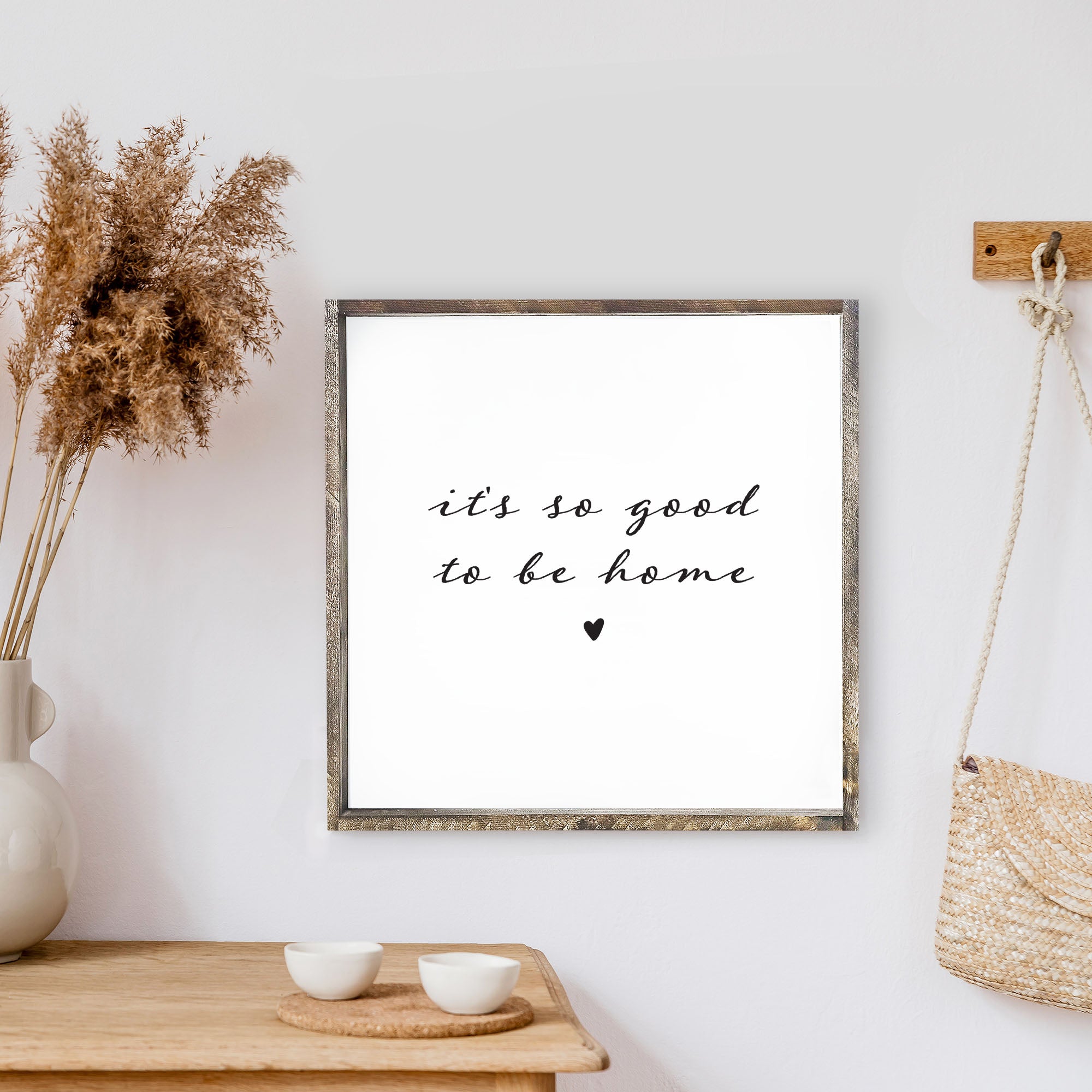 A charming 13x13 inch wood sign with the phrase 'It's So Good To Be Home' in black text on a white background, framed in stained pine wood.