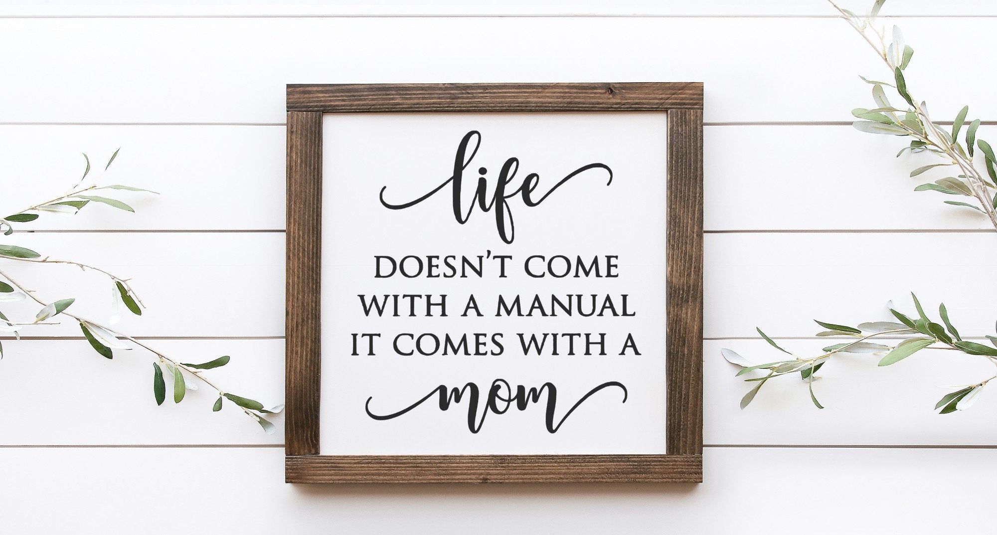 A handmade wooden sign with the phrase 'Life Doesn't Come With A Manual It Comes With A Mom' painted in elegant lettering on a matte white background.