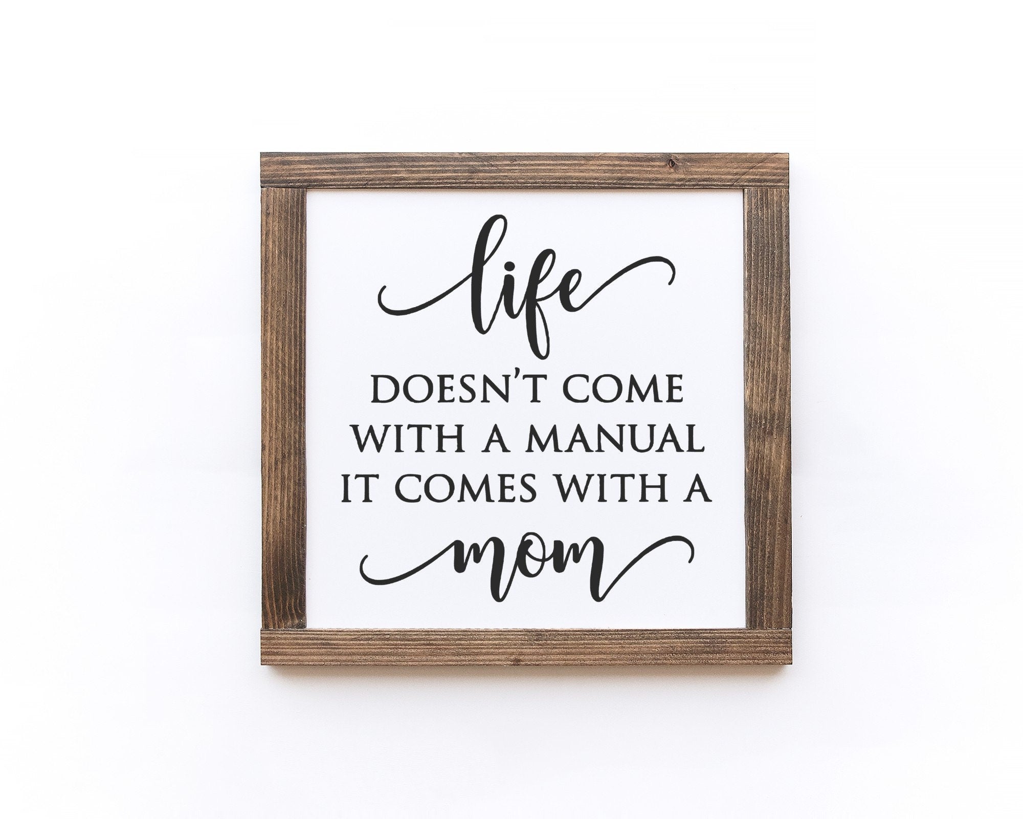 A handmade wooden sign with the phrase 'Life Doesn't Come With A Manual It Comes With A Mom' painted in elegant lettering on a matte white background.