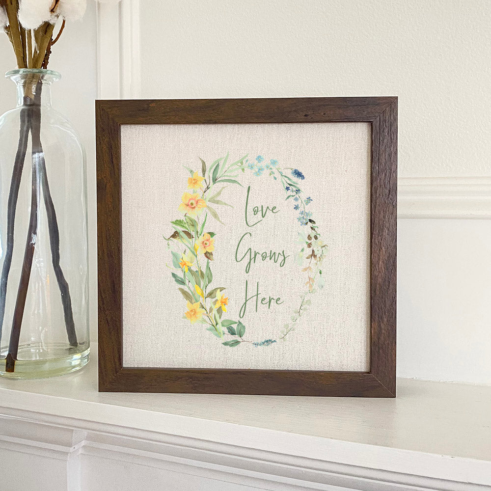 Love Grows Here Wreath framed sign with a stylized wood frame, eco-friendly printing, and a linen-look neutral background.