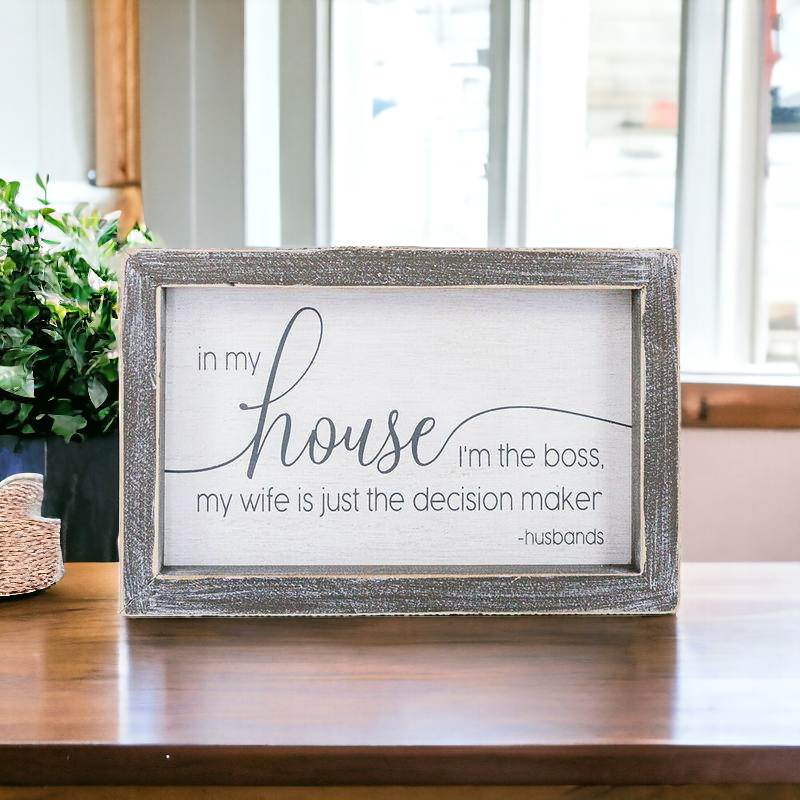 A humorous wooden sign that reads 'In My House I'm The Boss, My Wife Is Just The Decision Maker - Husbands', framed in white and grey.