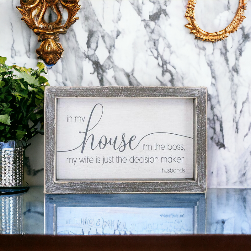 A humorous wooden sign that reads 'In My House I'm The Boss, My Wife Is Just The Decision Maker - Husbands', framed in white and grey.