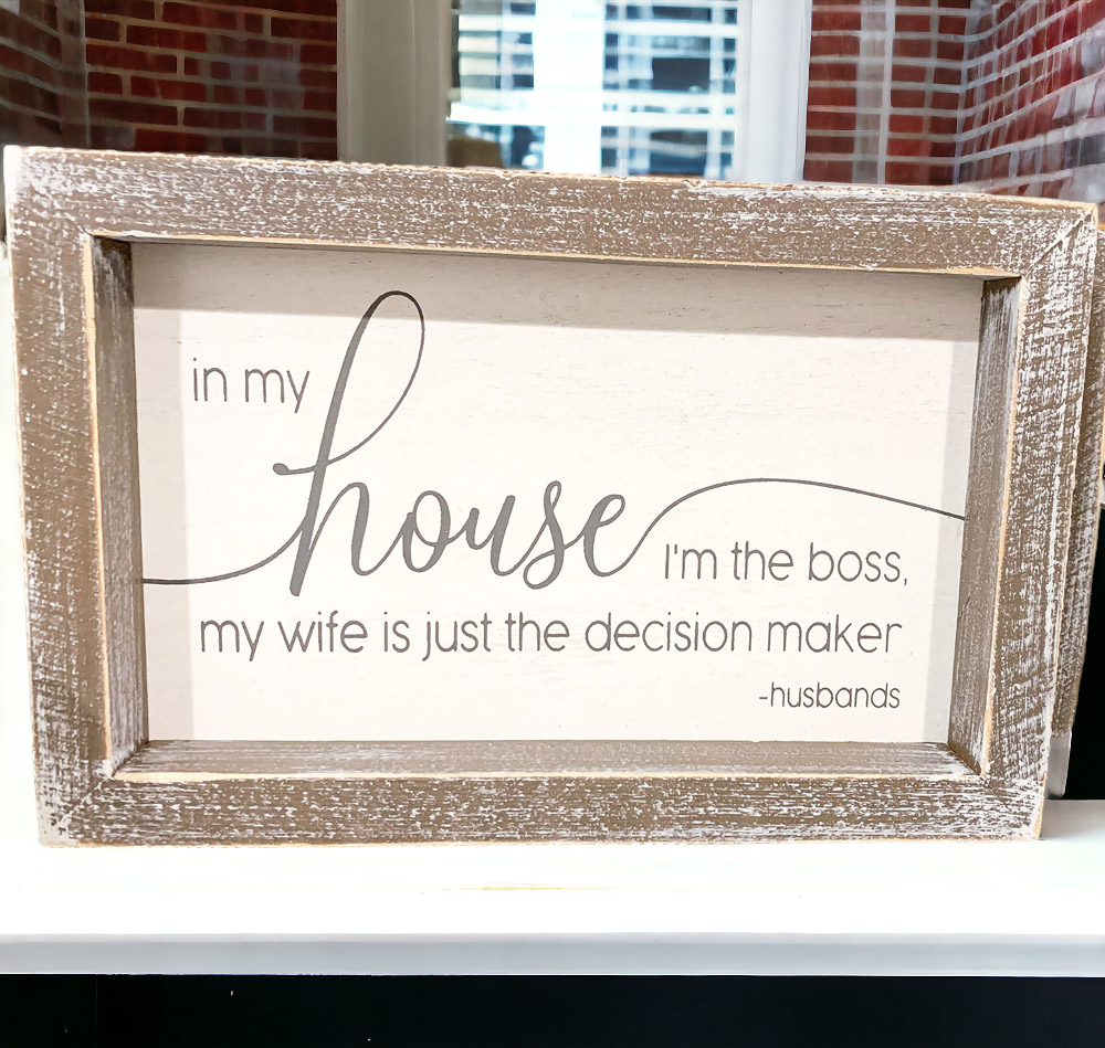 A humorous wooden sign that reads 'In My House I'm The Boss, My Wife Is Just The Decision Maker - Husbands', framed in white and grey.